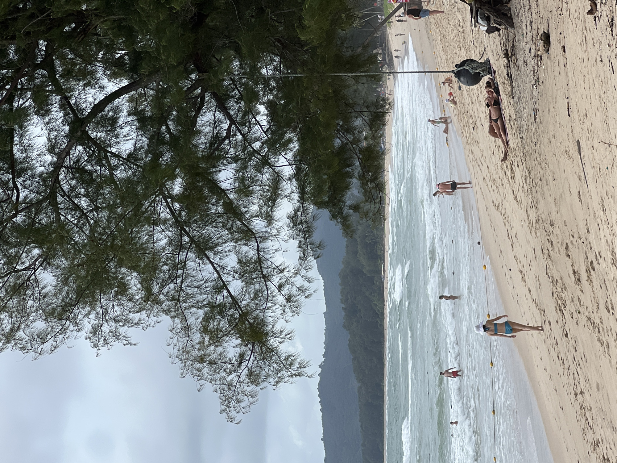 This is a medium-shot, eye-level, vertical photograph taken at Bang Tao Beach in Choeng Thale, Thailand. The image captures a cloudy day at a tropical beach. In the foreground, a large, leafy pine tree casts a shadow over the sandy beach. Two people are lying on their backs on the sand, close together, under the tree. Further up the beach, several other individuals are visible, some walking and some lying on towels. In the middle ground, the ocean has moderate waves, and several people are in the water, some wading and others swimming. A yellow rope with buoys marks a boundary in the shallow water. In the background, a lush green hillside covered in trees rises above the beach, and the sky is overcast. The overall scene depicts a relaxed atmosphere at a popular beach destination.