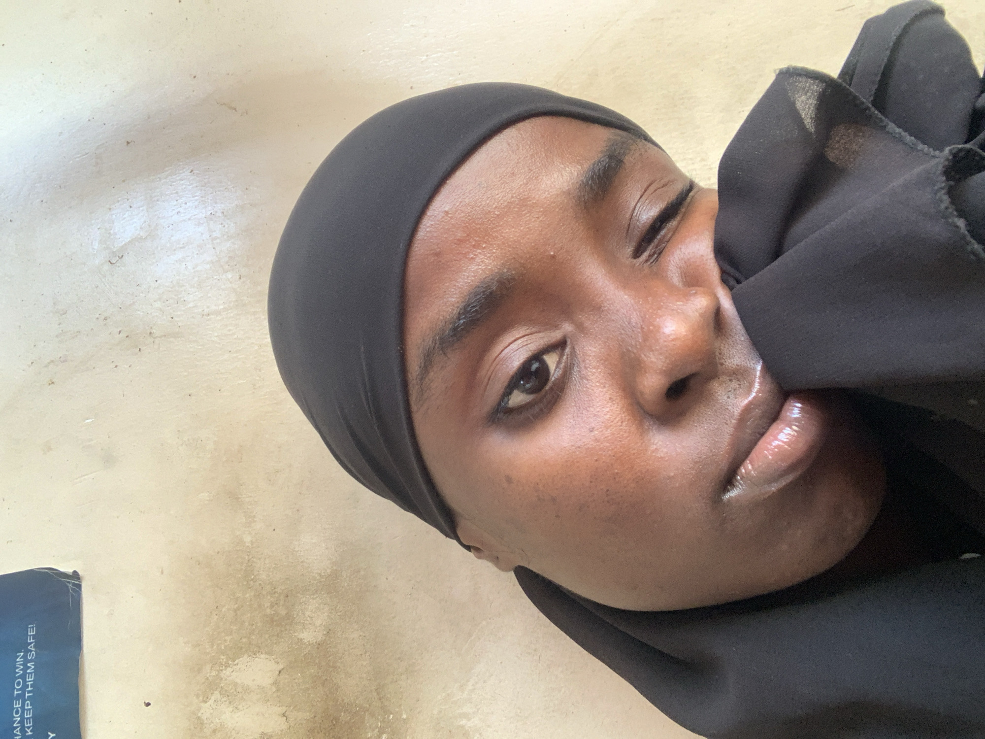 A medium shot depicts a Black woman wearing a black headscarf. She is positioned against a light, slightly textured surface suggestive of flooring. The woman's face, angled towards the camera, shows a partially obscured left eye, and she appears to be holding a piece of her scarf to her face. The lips are visibly glossy. The woman is likely inside based on the flooring, but no other spatial clues provide information about the city of Adankolo, Nigeria. A partially visible brochure in the lower left corner. It contains the partially visible words "chance to win" and "keep them safe!". The scene is static.