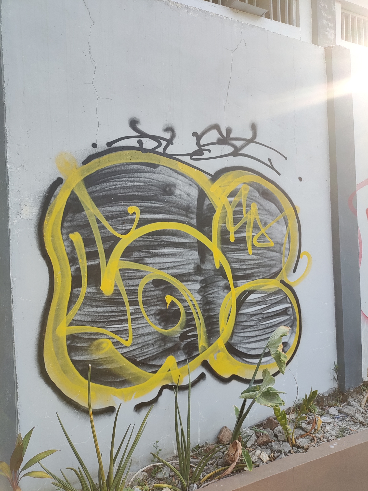 The image depicts graffiti on a light gray concrete wall. The graffiti features a large, yellow, bubbly outline surrounding a black and white abstract design with horizontal lines. Above the main graffiti, there are two smaller black spray-painted tags. To the right of the wall, a bright sunbeam is visible, suggesting it is daytime and possibly late afternoon due to the angle of the light. The wall appears to be outdoors, as indicated by the presence of plants and rocks at its base. The specific location in Cirebon, Indonesia, is not discernible from the image alone.