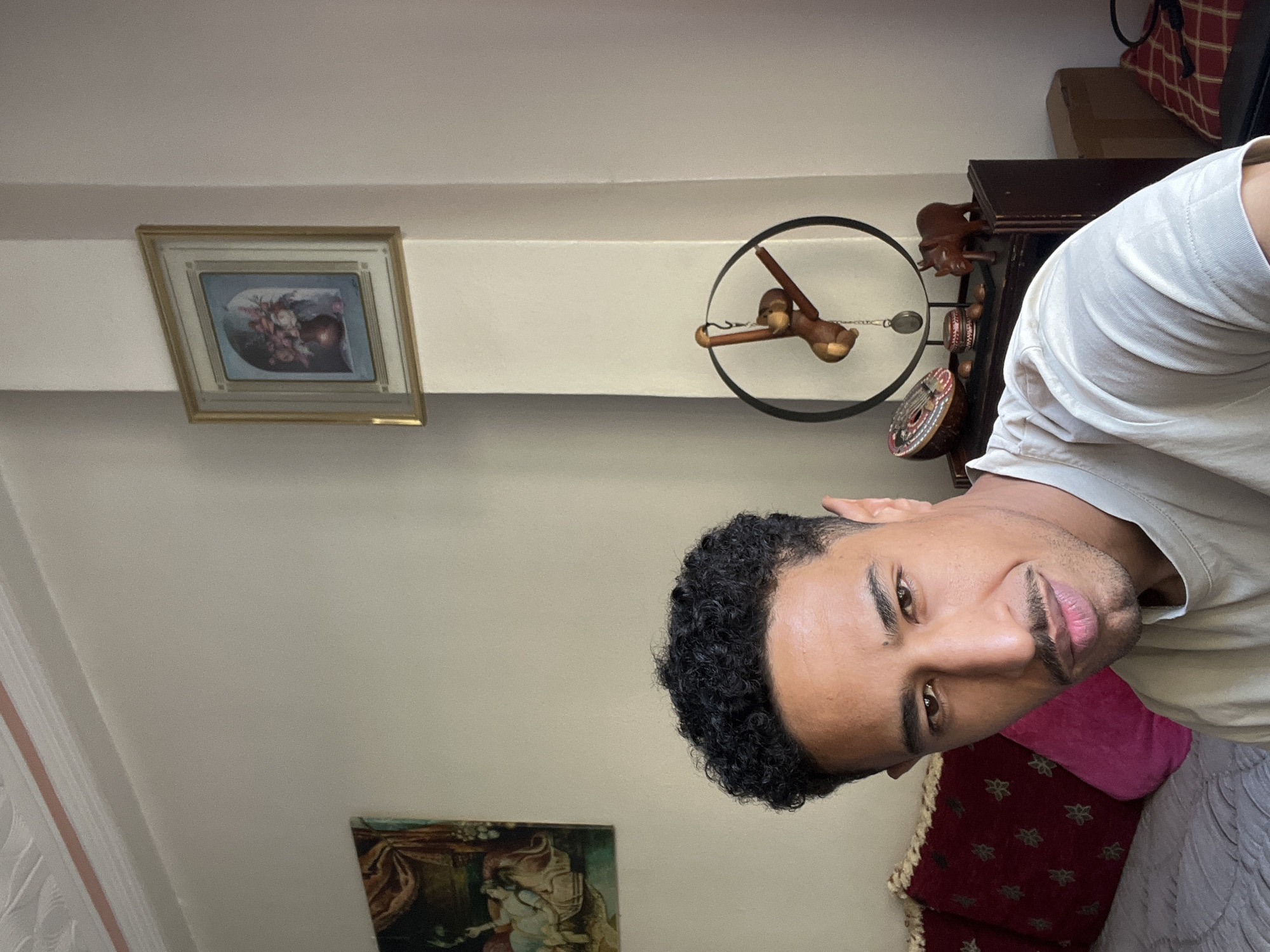 A medium shot depicts a man with dark curly hair, light brown skin, and a short beard, wearing a white t-shirt. He appears to be indoors, likely in a residence in Tanger طنجة, Morocco, based on the interior design. Behind him is a wall with two framed paintings, one above the other. The upper painting has a gold frame and appears to be a floral scene. The lower painting depicts people and a landscape. To the right, there's a dark wooden piece of furniture with what looks like a pendulum clock and a decorative monkey figure. Red pillows are seen behind him on the right, and the edge of what seems to be a grey bedspread is also visible. The room has white walls.