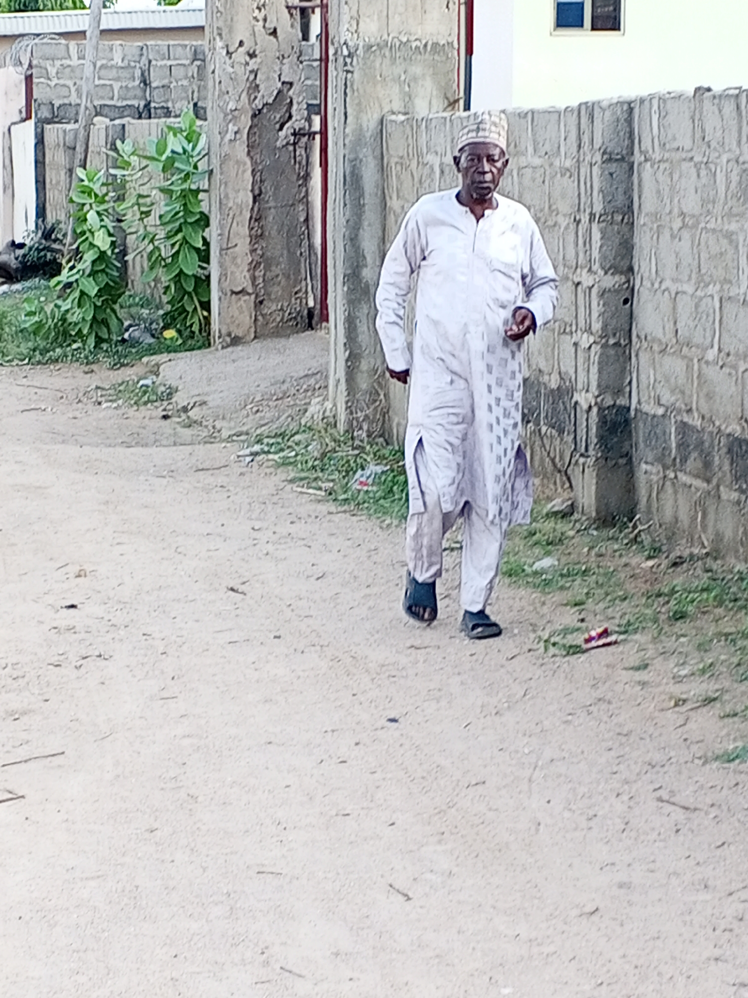 An elderly man walks on a dirt road in Adonkolo, Nigeria. He wears a light-colored, patterned robe and a cap, with dark sandals on his feet. To his right is a concrete block wall. To his left, the corner of a building and some plants can be seen. The road is light in color and appears unpaved.