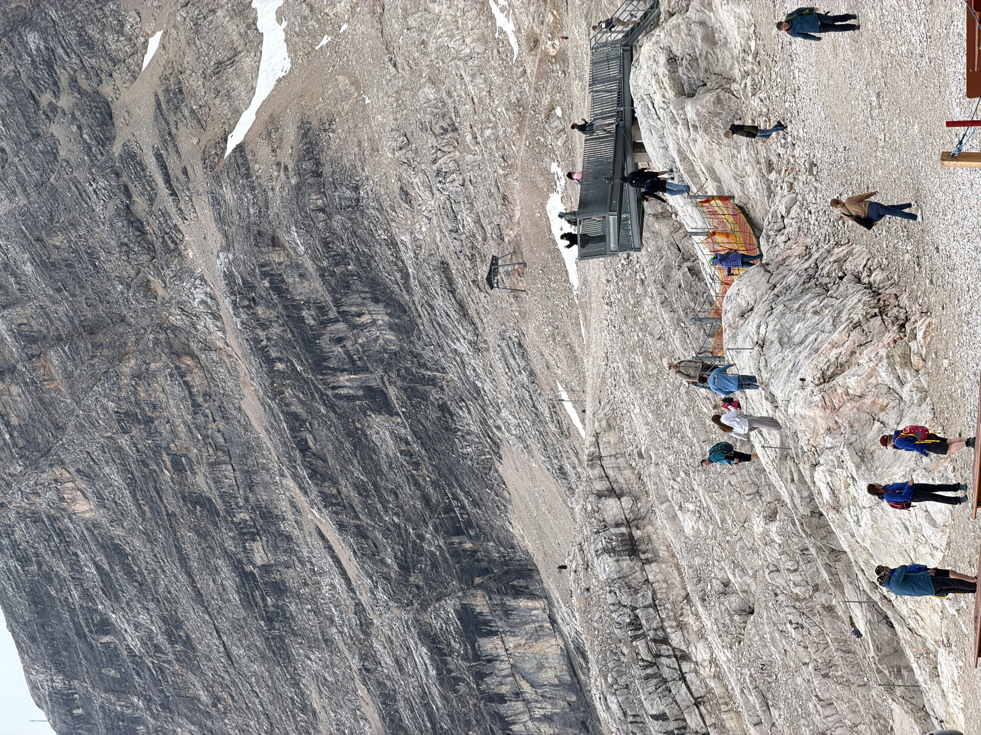 This image depicts the Zugspitze, Germany's highest peak, located near Garmisch-Partenkirchen. The scene is set on a rocky mountainside, with a significant portion of the frame occupied by the imposing, grey, stratified rock face of the mountain. Several groups of tourists are visible scattered across the rocky terrain and on a metal observation platform.
On the platform, which extends out over the slope, a number of individuals are standing, some looking out at the view. To the left of the platform, a group of people are navigating the rocky ground, some walking and others standing. Further down the slope, more individuals are walking along paths and amongst the rocks. One person in a dark outfit stands on a prominent rock formation in the mid-ground. In the background, a small wooden structure on stilts is visible, possibly an observation point or equipment shed.
The environment is characterized by rough, uneven, grey and beige rocks, with patches of lingering snow or ice visible higher up the mountain. Safety railings and guide ropes are present in some areas to assist hikers and visitors on the steep terrain. The overall atmosphere suggests a popular tourist destination with ample opportunities for sightseeing and hiking.