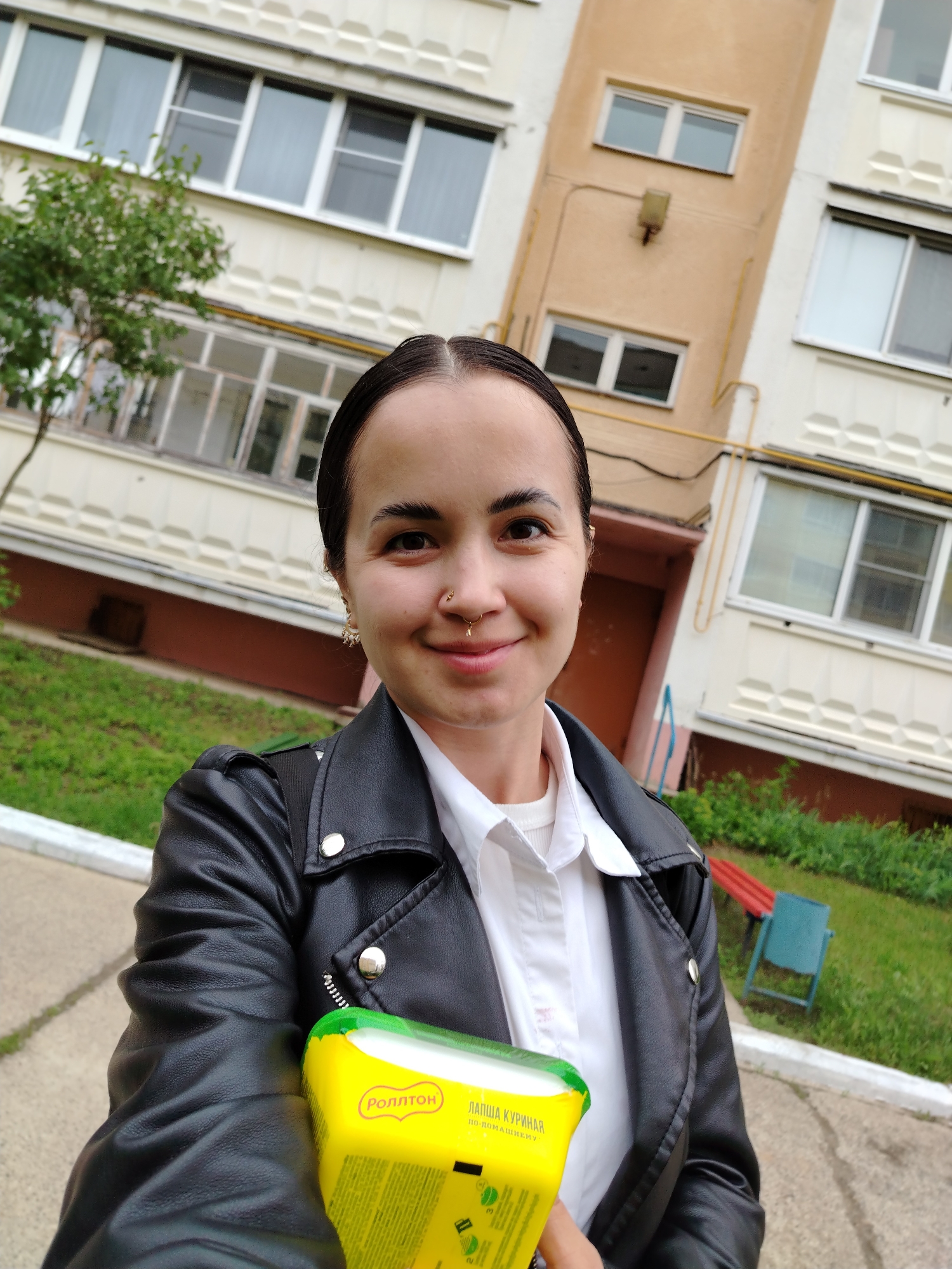 A young adult female is depicted in a selfie, taken outdoors in Noviy, Russia. She has dark hair pulled back, a pale complexion, dark eyebrows, and dark eyes. She displays a slight smile. Visible facial piercings include a gold hoop in her left nostril and a gold septum ring. She wears dangle earrings, with one visible on her left ear. Her attire consists of a white collared shirt over a white ribbed undershirt, beneath a black leather-style jacket featuring silver snap buttons and a zipper detail. She holds a yellow rectangular container with a green top in her left hand, angled towards the camera. Cyrillic text on the container identifies it as "РОЛЛТОН" (Rollton) brand "ЛАПША КУРИНАЯ ПО-ДОМАШНЕМУ" (Chicken Noodles Homemade style).
The background features two multi-story residential buildings. The building to the viewer's left is light-colored, possibly cream, with white-framed windows and decorative patterned panels below the windows. Enclosed balconies are visible on its lower floors. To the viewer's right, a distinct light brown/beige building with white-framed windows and yellow exterior gas piping is present, along with a brown door at ground level and an exterior light fixture. A small green leafy tree is situated to the left foreground. The ground consists of a concrete or paved pathway with a white curb, bordered by patches of green grass. In the far right background, a red and blue bench-like structure and a blue trash can are visible in a grassy area. The sky appears overcast.
