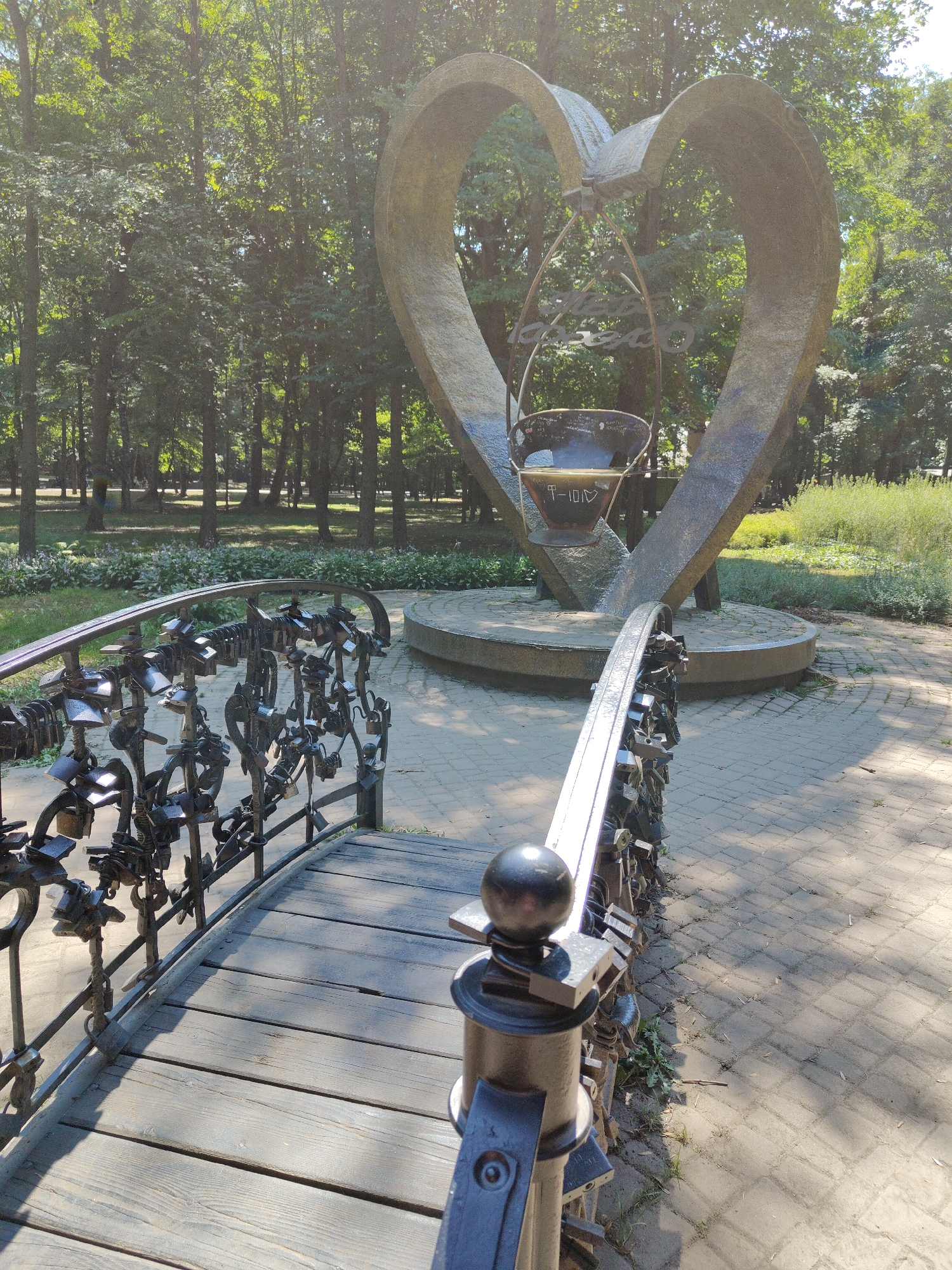 The image depicts a large, heart-shaped sculpture in a park setting, likely in Chernivtsi, Ukraine, given its common association with the city's symbolism. In the foreground, a pedestrian bridge with a wooden deck and a metal railing covered in numerous padlocks is visible. The railing itself is intricate, with decorative metalwork and many padlocks attached, suggesting a tradition of couples locking their love. Behind the bridge, the heart sculpture stands prominently. It is a large, metallic structure, open in the center, and appears to have a suspended element within it, possibly a smaller receptacle or decorative piece. The sculpture is situated on a circular concrete base. The park environment is characterized by lush green trees forming a dense backdrop, with sunlight filtering through the leaves, creating dappled shadows on the ground. The pathway around the sculpture is paved with light-colored bricks. The overall scene conveys a romantic and tranquil atmosphere, common for public art installations dedicated to love and relationships.