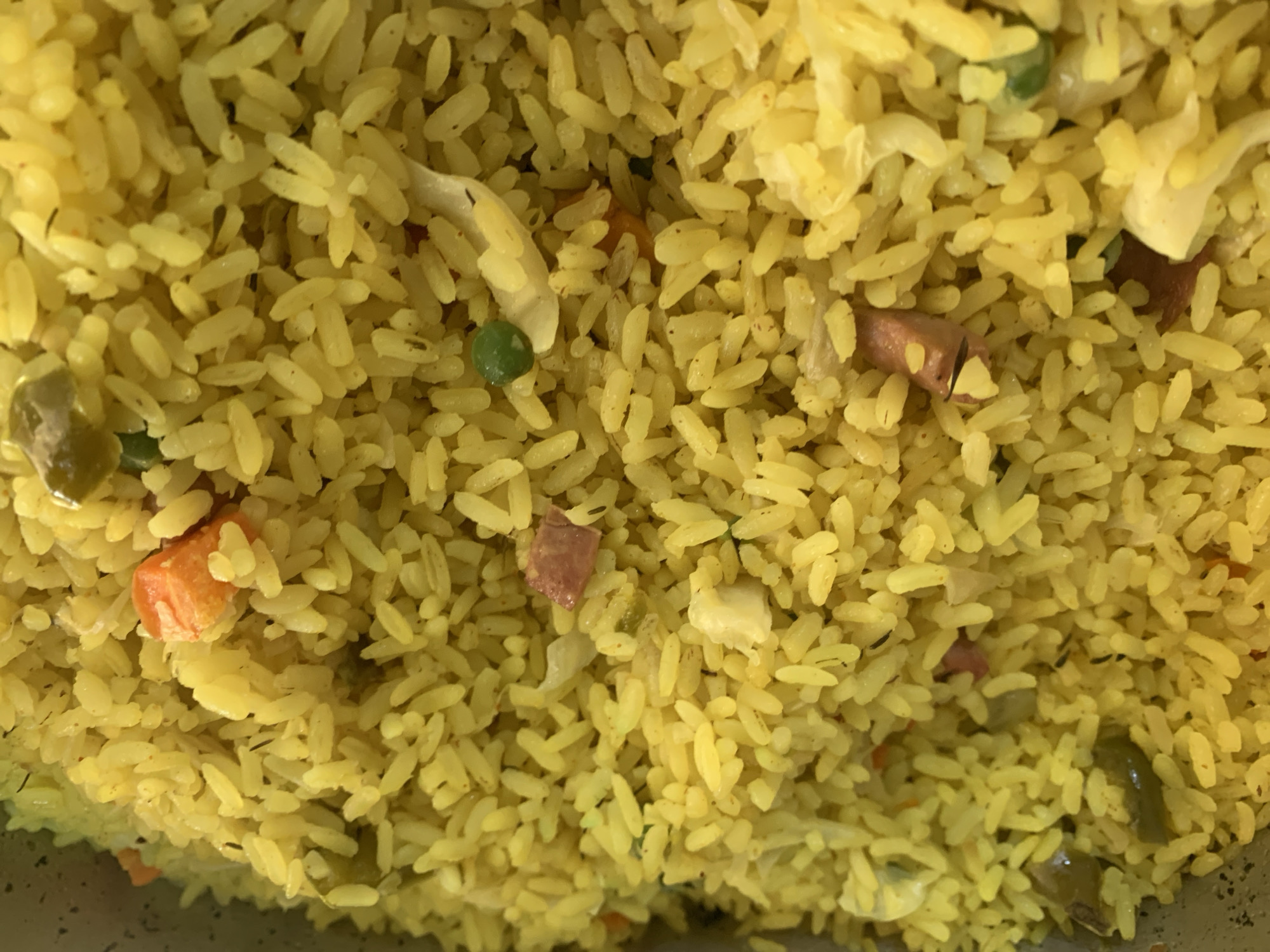 A close-up, top-down view of a large portion of yellow fried rice. The rice is interspersed with pieces of diced carrots, peas, and small chunks of ham. The dish is likely served in a large metal container, as indicated by a sliver of silver metal visible at the top left of the frame. The lighting appears to be natural or overhead ambient light, creating a warm and slightly diffused illumination across the entire surface of the food. The texture of the rice is granular, with individual grains visible. The overall impression is one of a plentiful serving of a savory dish.