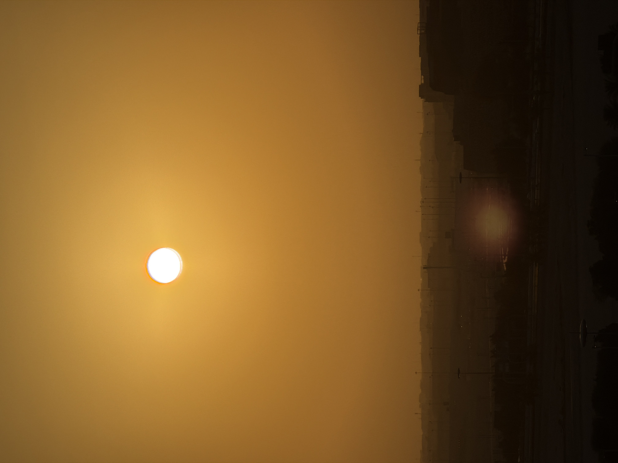 This media file captures a scene dominated by a hazy, golden sky, suggesting either sunrise or sunset. A bright, white sun is prominently positioned in the left half of the frame, its light diffused by atmospheric conditions. To the right, the silhouette of a city skyline is visible, with buildings appearing as dark shapes against the muted sky. A highway or road runs through the scene, marked by streetlights that faintly glow. A lens flare, a bright reddish-orange orb, is present in the right half of the image, interacting with the sunlight and the atmospheric haze. The overall impression is one of a quiet, perhaps serene, urban landscape at dusk or dawn, with the weather contributing to a softened, almost dreamlike quality. No people are discernible in the image, and no text is visible.