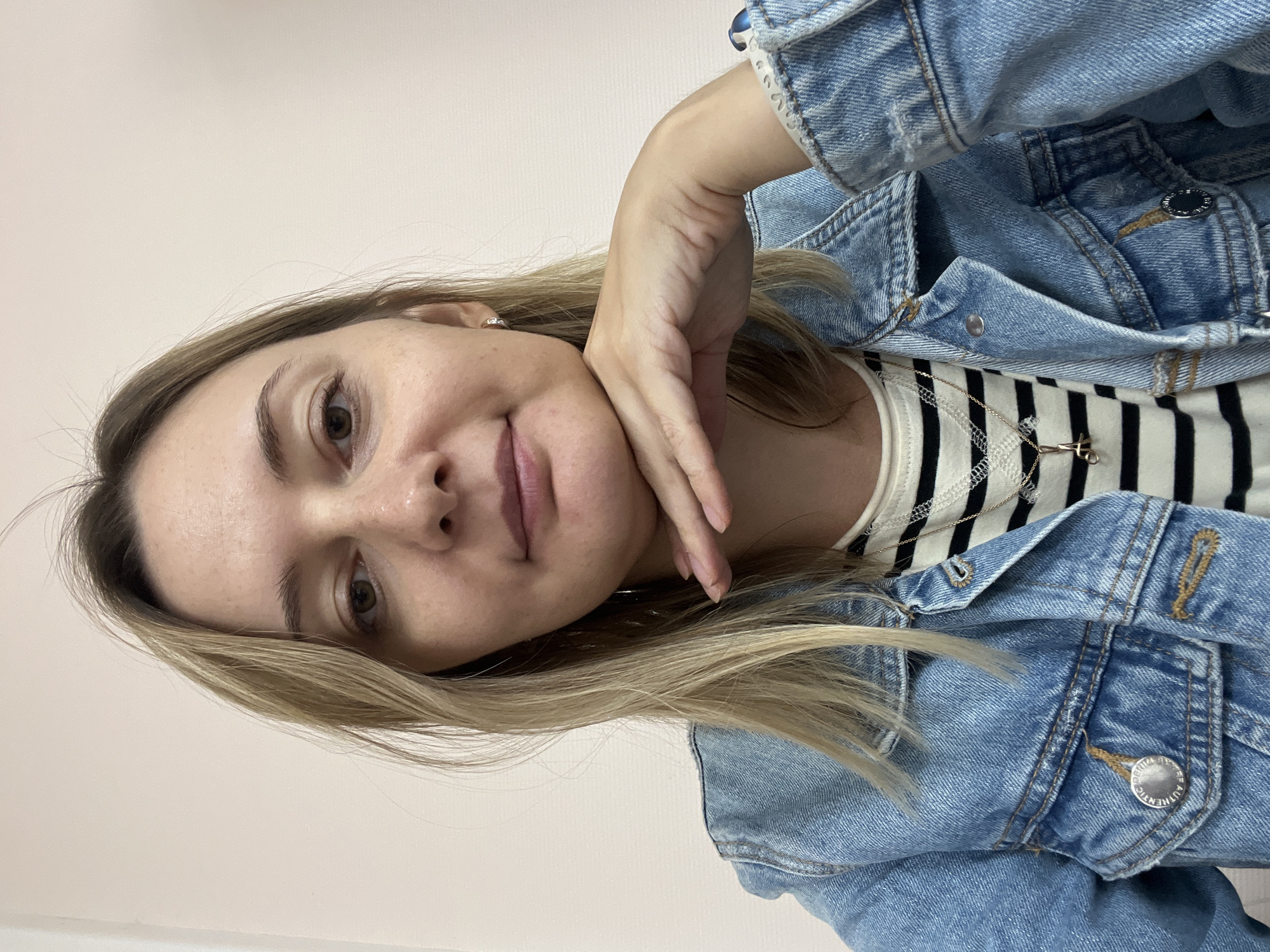 A medium close-up shot shows a light-skinned woman with shoulder-length blonde hair. She is wearing a blue denim jacket over a black and white striped shirt. A delicate gold chain with a small pendant hangs around her neck. Her right hand is resting on her chin, with her fingers gently touching her cheek. The background is a plain, light-colored wall. The woman is looking directly at the camera with a neutral expression. There is no indication of the city of Volgograd, Russia, in this image.