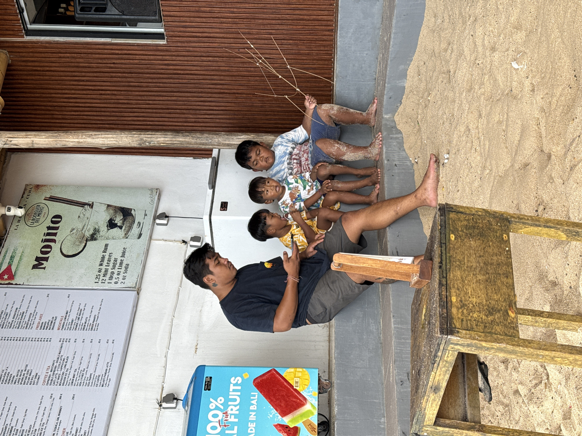 A medium shot captures a man and three children, likely in Kuta Selatan, Indonesia, based on the "MADE IN BALI" text visible. The man, dark-haired and wearing a dark t-shirt and shorts, sits on the ground, seemingly interacting with a child on his lap. Three children sit behind him, sandy-legged. The child at the top is holding what appear to be dried branches.
To the left is a menu board advertising "Mojito" and other beverages, near an ice cream freezer displaying "100% ALL FRUITS." Behind the man and the children, there is beach sand. A wooden structure, possibly a bench or table, is on the right of the frame. A corrugated metal wall is in the background, above the menu board. The scene appears to be an outdoor beachfront or near-beach location.