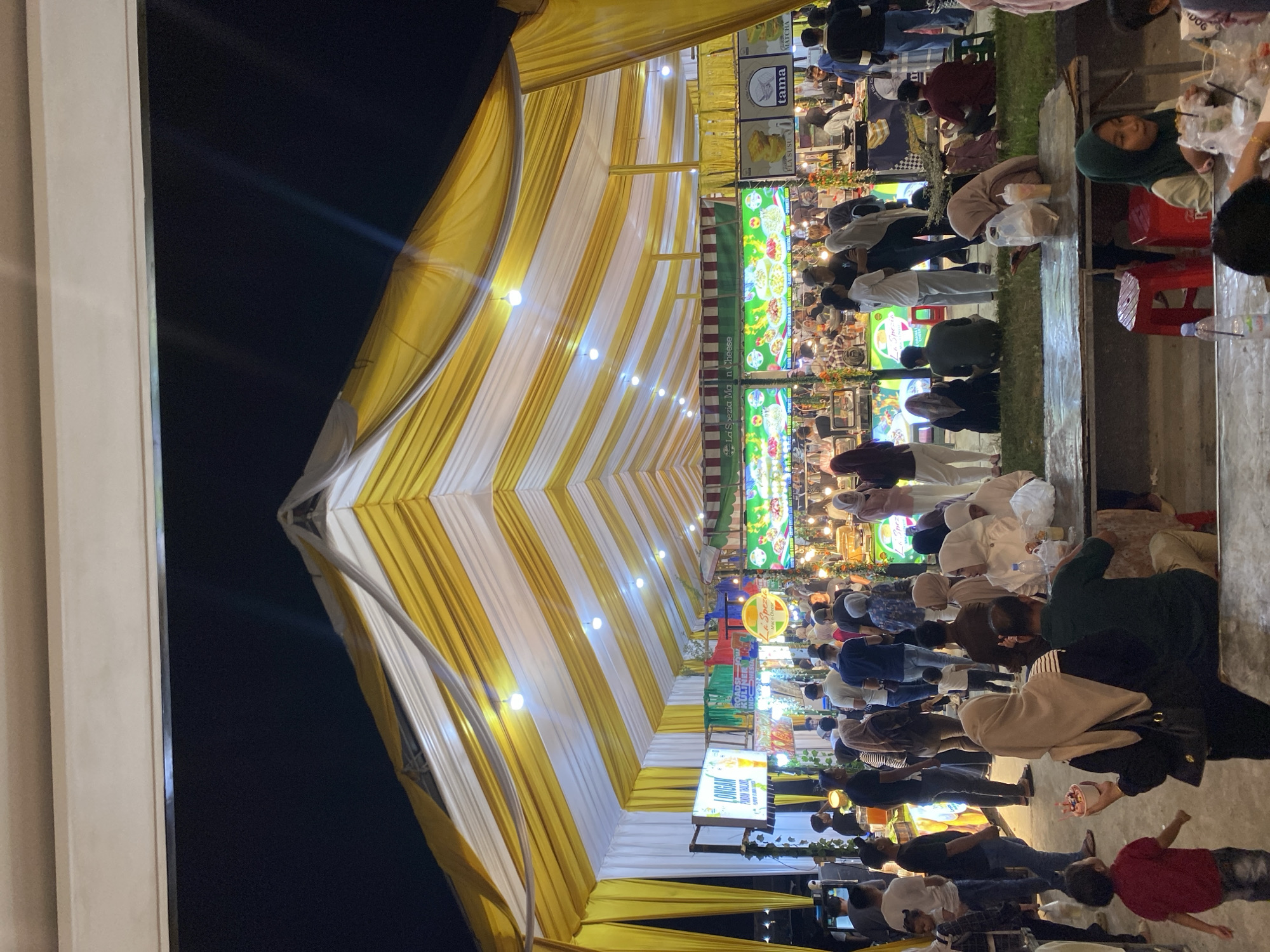 This media file depicts a lively night scene at a food market in Lhokseumawe, Indonesia. The focal point is a large, striped yellow and white tent structure, brightly lit by small bulbs, creating a festive atmosphere. Beneath the tent, numerous people are gathered, suggesting a bustling event or a popular marketplace.
The crowd is diverse, with individuals of various ages and genders, many wearing casual attire. Some people are seen queuing or standing at food stalls, while others are seated, enjoying their meals. The presence of food stalls, indicated by the visible signage and the nature of the gathering, points to a culinary event.
Several signs with text are visible, including "PANDAN THAILAND", "La Spezia Man Cheese", "FLA SUSU", and "MATCHA". These likely represent food or drink items being sold. The overall mood appears energetic and convivial, characteristic of a night market or a local festival.
The lighting suggests that the event is taking place after sunset, with artificial lights illuminating the scene. The ground appears to be paved, consistent with an urban market setting. The image captures the vibrancy and social interaction typical of such gatherings in Indonesia.