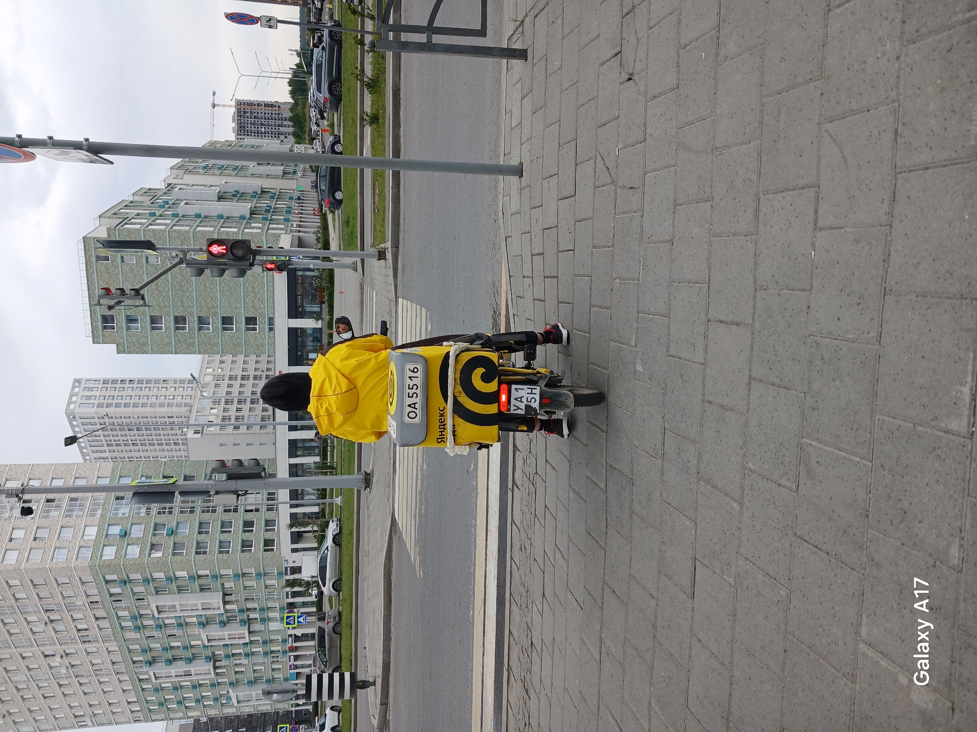 An image taken in an urban environment in Yekaterinburg, Russia, depicts a delivery person on a yellow scooter stopped at a red traffic light. The person is wearing a bright yellow jacket and a black helmet, with a large yellow delivery bag or box adorned with the "Яндекс" (Yandex) logo. The number "OA 5516" is visible on the delivery box, and the scooter's license plate shows "YA1 5H."
The scooter is positioned on the road, adjacent to a grey paved sidewalk. A pedestrian crossing is marked on the asphalt. The traffic light, visible to the left, displays a red signal for pedestrians (a red standing figure) and vehicles, indicating a stop. In the background, modern multi-story residential buildings with numerous windows line the street; one building features a distinctive light green facade. Further in the distance, a construction crane and a building under construction are visible, suggesting urban development. Parked cars are seen along the left side of the road.
The sky is overcast, indicating daytime conditions. The image includes various street signs and poles. A "Galaxy A17" watermark is visible in the bottom right corner.