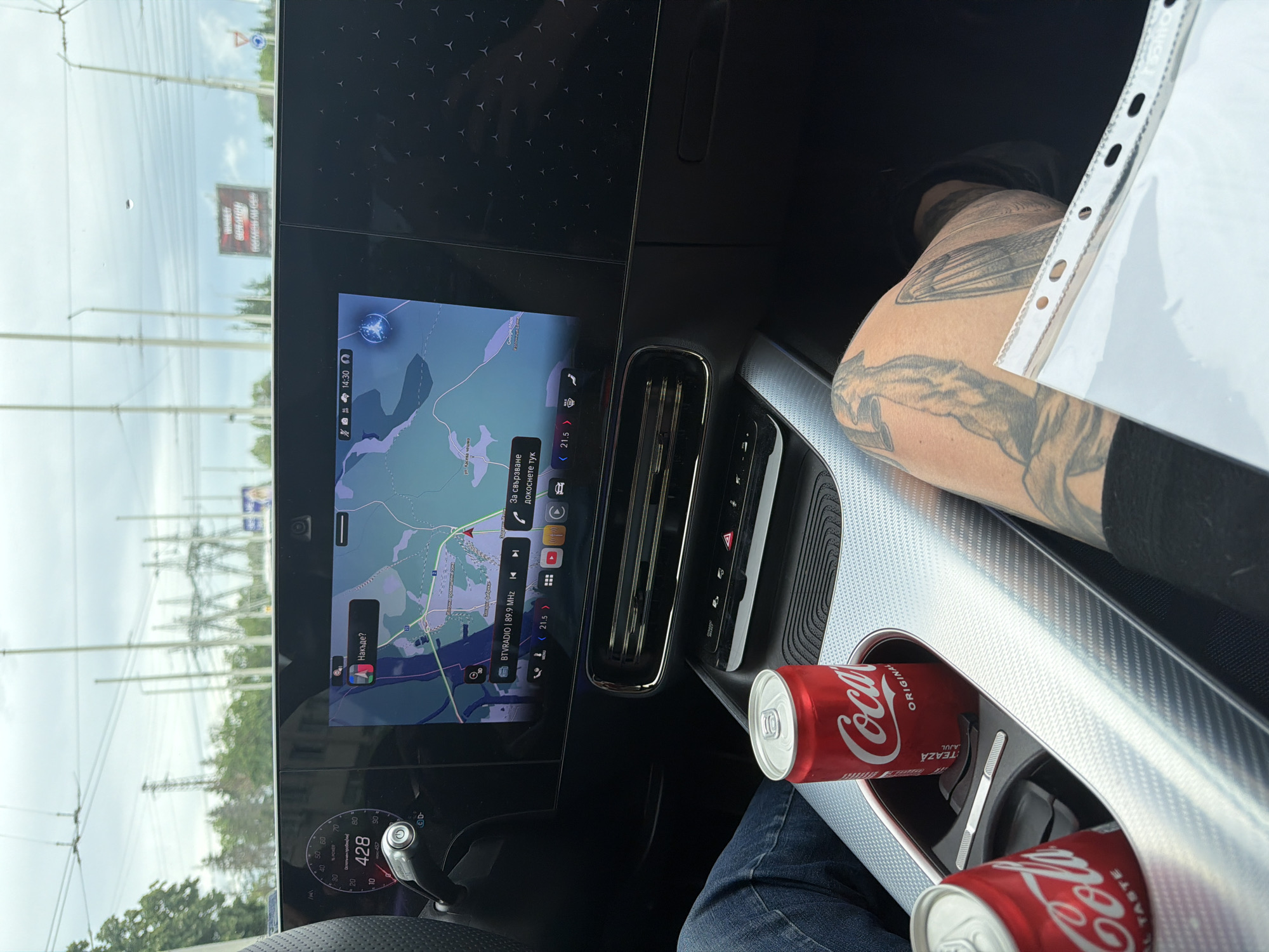 The image captures the modern interior of a car, likely a Mercedes-Benz given the dashboard design and star-patterned trim, viewed from the passenger's perspective towards the driver's side and the road ahead.
**Subjects:** The central focus is the car's sophisticated digital dashboard, featuring two large screens. On the right, a tattooed arm and a leg clad in blue jeans are visible, belonging to a person resting in the passenger seat. Two red Coca-Cola Original Taste cans are nestled in the cup holders of the car's sleek center console, which is accented with a textured silver trim.
**Scene and Setting:** Through the windshield, an overcast sky is visible, along with utility poles, power lines, and trees, suggesting an outdoor, possibly highway or main road setting. A distant, blurred billboard is also present. The scene depicts travel within or around Ruse, Bulgaria, as indicated by the context and the Cyrillic text on the navigation screen.
**Activity or Event:** The car is either in motion or stopped briefly during a journey. The active Google Maps navigation on the main screen indicates travel is underway.
**Notable Details:**
* **Car Features:** The digital screens display information such as the time "14:30", the temperature "< 21.5 >", and "BTVRADIO | 89.9 MHz". The instrument cluster shows "428" (likely mileage or range) and a "POWER" indicator.
* **Navigation:** Google Maps shows a map with Cyrillic place names, and prompts like "Накъде?" (Where to?) and "За свързване докоснете тук" (To connect, tap here).
* **Person's Details:** The visible arm features intricate tattoos, including what appears to be a fox and possibly other design elements. A clear plastic document holder is resting on the person's lap.
* **Atmosphere:** The cloudy sky suggests a mild or potentially rainy day.
**Visible Text:** "Coca-Cola ORIGINAL TASTE", "14:30", "428", "POWER", "Накъде?", "За свързване докоснете тук", "Google Maps", "BTVRADIO | 89.9 MHz", "< 21.5 >".