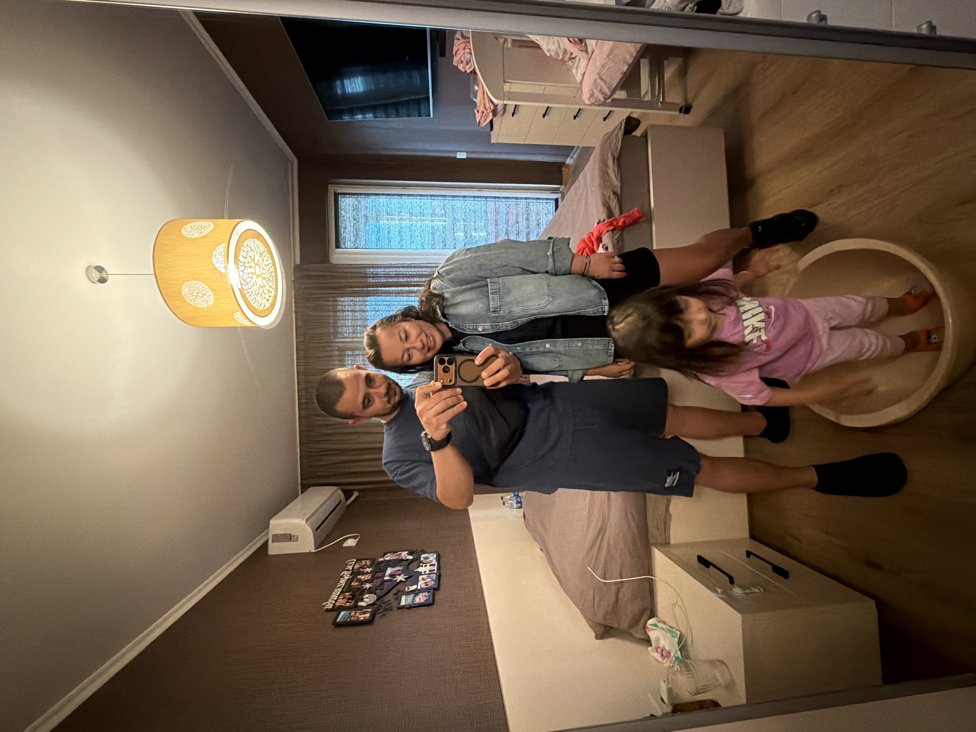A family of three, two adults and a young child, are taking a mirror selfie in what appears to be a bedroom. The adults, a man and a woman, are standing and smiling while holding a phone to capture the image. The man is wearing a dark blue t-shirt and shorts, and the woman is wearing a denim jacket over a dark top and shorts. The young child, a girl, is standing in a large, round basin. She is wearing a pink t-shirt and pink pants. The room has a brown textured wallpaper, a bed with a brown duvet, bedside tables, and a ceiling lamp. There's also an air conditioner mounted on the wall and a television. The overall impression is a casual, domestic setting, likely within an apartment or house. The context of Burgas, Bulgaria, suggests this might be their home.