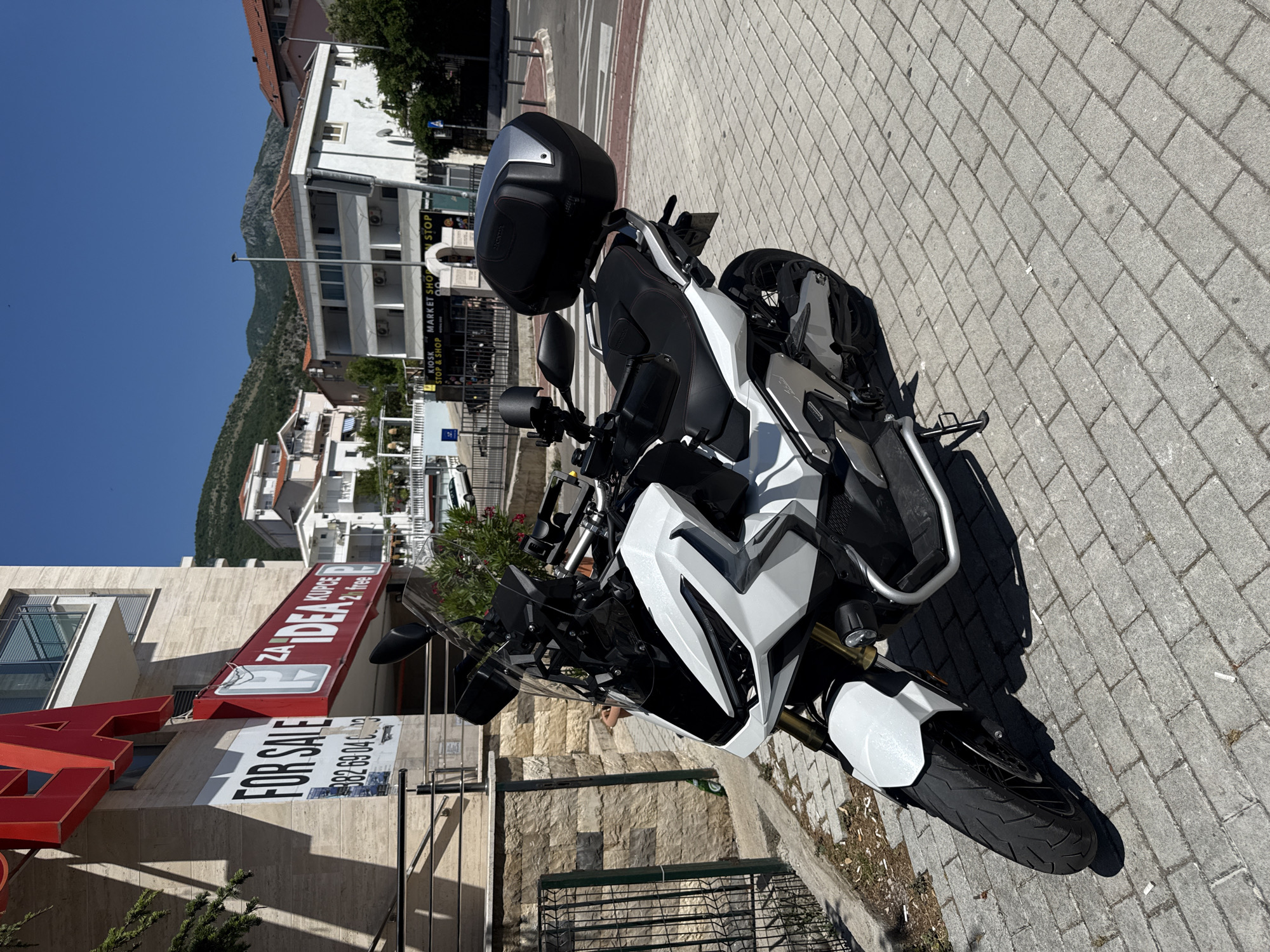 A white and black Honda ADV 750 scooter is parked on a light gray brick-paved sidewalk in Rafailovići, Montenegro, under a clear blue sky. The scooter, facing forward and slightly to the left, features a prominent windscreen, golden front forks, and a black top case with silver accents bearing the Honda logo. It rests on its side stand. To the left, a multi-story building with a light-colored facade displays large red signage including "ZA IDEA KUPCE P 2h FREE" (For Idea customers, parking 2 hours free) and a white "FOR SALE" sign with a partially visible phone number "082 69 04 33". Further into the scene, across a paved road with a visible crosswalk and a small intersection, stands a white building with a red-tiled roof, featuring multiple exterior air conditioning units and signs reading "KIOSK STOP & SHOP" and "MARKET SHOP". Green, rocky mountains are visible in the distant background. The strong sunlight casts distinct shadows, indicating a bright day. The sidewalk has minimal debris, including small white fragments that resemble cigarette butts. No individuals or other vehicles are present.