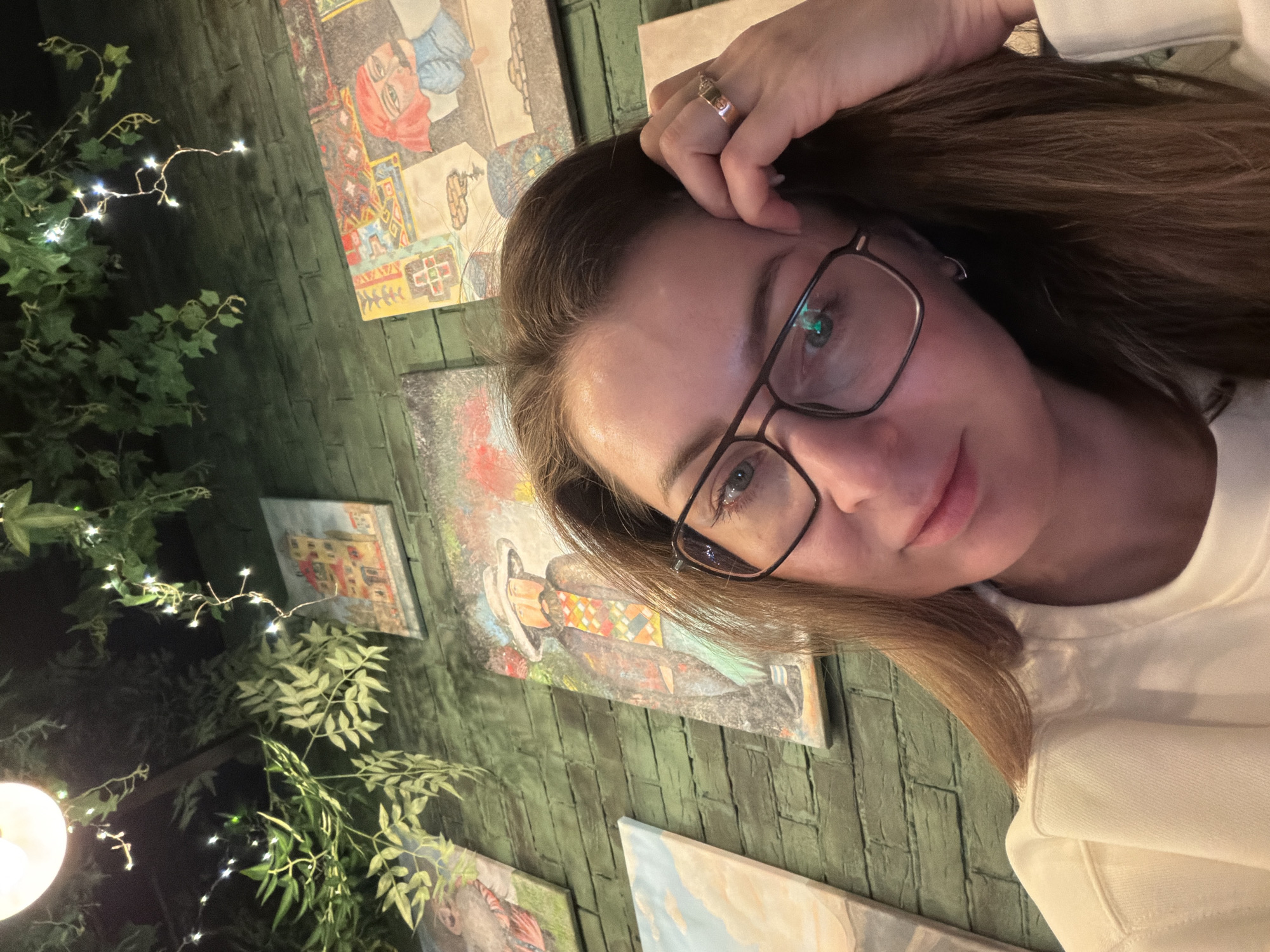 A woman with light brown hair and wearing glasses poses for a selfie. Her right hand is touching her forehead, and she is wearing a ring on her right ring finger. She is wearing a light-colored sweater. The background features a green brick wall adorned with several paintings, and green leafy plants with string lights. Based on the paintings, the location is possibly a cafe or art space in Izhevsk, Russia. The image is rotated counter-clockwise.