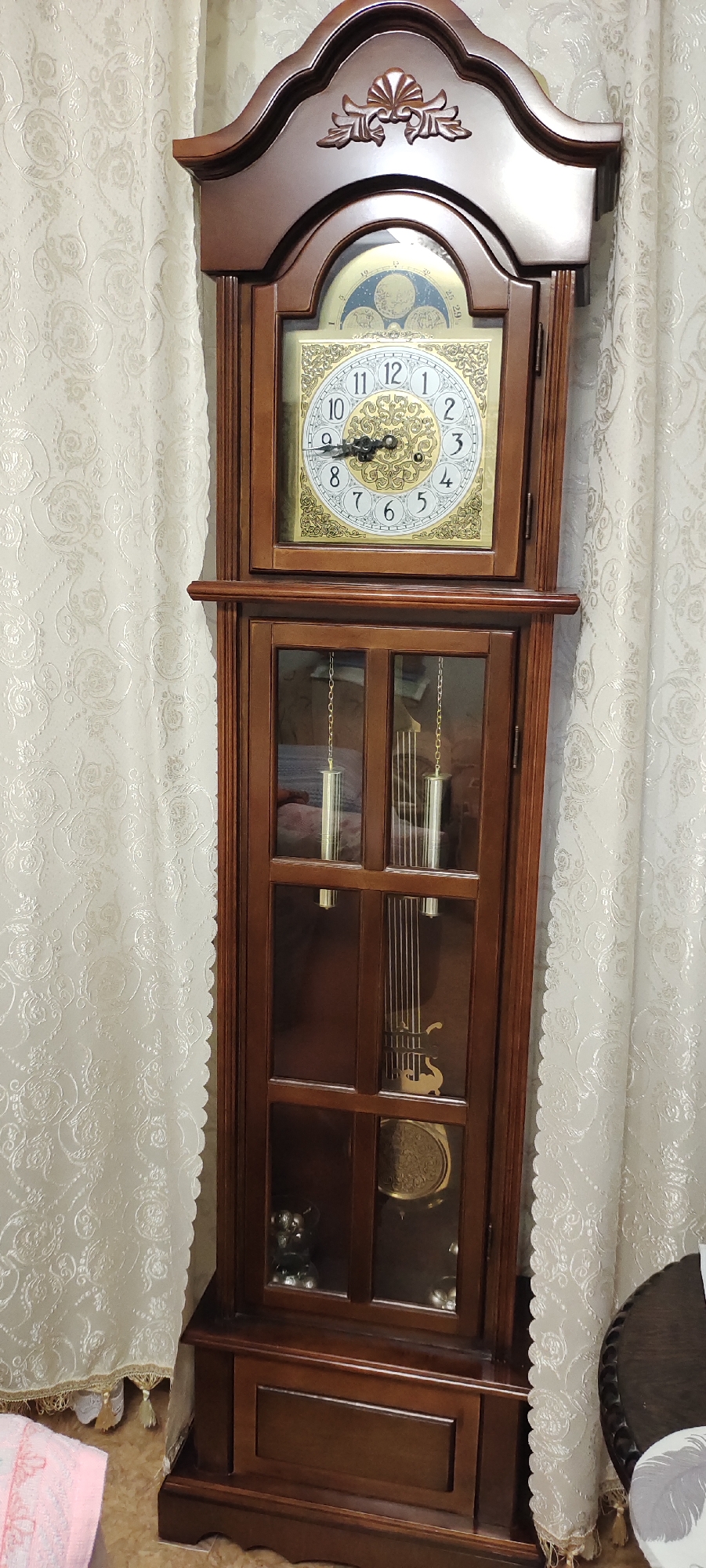 This media file is a full-shot, vertically oriented image of a grandfather clock. The clock is made of dark wood and features a decorative top with carved detailing. Below the top, a glass-enclosed section reveals the clock's face and pendulum. The clock face is round with Roman numerals and ornate gold detailing. The hour and minute hands are black and appear to be set at approximately 9:12. Above the main clock face is a smaller dial, likely for moon phases or other complications, with a blue background and celestial imagery.
The clock has a glass-doored cabinet in the lower section, revealing hanging weights and the pendulum. The pendulum itself is golden and has a decorative disc at its base, showing intricate carvings. Visible within the cabinet are some decorative objects, appearing to be small metallic figurines and a larger round decorative element.
The grandfather clock is positioned indoors, in what appears to be a living room or hallway. It is set against a backdrop of cream-colored curtains with a subtle, ornate floral pattern. The lighting suggests it is daytime, but the weather is not discernible. There is no visible text in the image. To the left of the clock, a pink cloth is partially visible on the floor, and to the right, a small portion of a round, dark wooden table with carved edges is seen. The overall impression is of a well-maintained, traditional piece of furniture in a domestic setting.