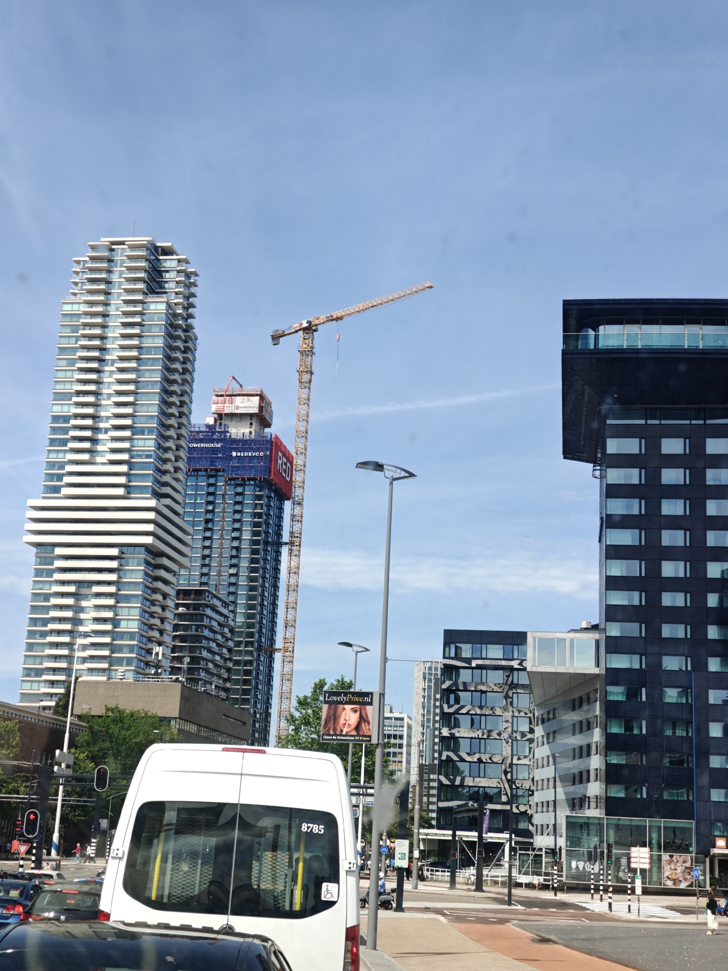 The image depicts a street scene in Rotterdam, Netherlands, likely near the Maas river, given the presence of modern high-rise buildings and construction cranes. In the foreground, a white van with the number 8785 is visible, indicating it is part of the local traffic. The van is stationary, suggesting a traffic jam or a stop at a traffic light. Several cars are behind the van, and a pedestrian can be seen in the distance.
The background is dominated by towering skyscrapers, characteristic of Rotterdam's contemporary architecture. One prominent building is a tall, white, multi-story structure with numerous balconies, suggesting a residential or mixed-use complex. Adjacent to it, another skyscraper is under construction, indicated by a large yellow crane and scaffolding. This building features a red banner with the text "RED" and "DREDVCO" and another banner with "OWERHOUSE". Further to the right, a dark, modern building with large windows and a cantilevered section stands out.
The sky is clear blue with a few wispy clouds. A streetlamp is positioned on the left side of the image, and traffic signals are visible, confirming the urban setting. A billboard advertising "LovelyPrive.nl" with an image of a woman is also present. The overall impression is one of a bustling city environment with ongoing development and modern infrastructure.
