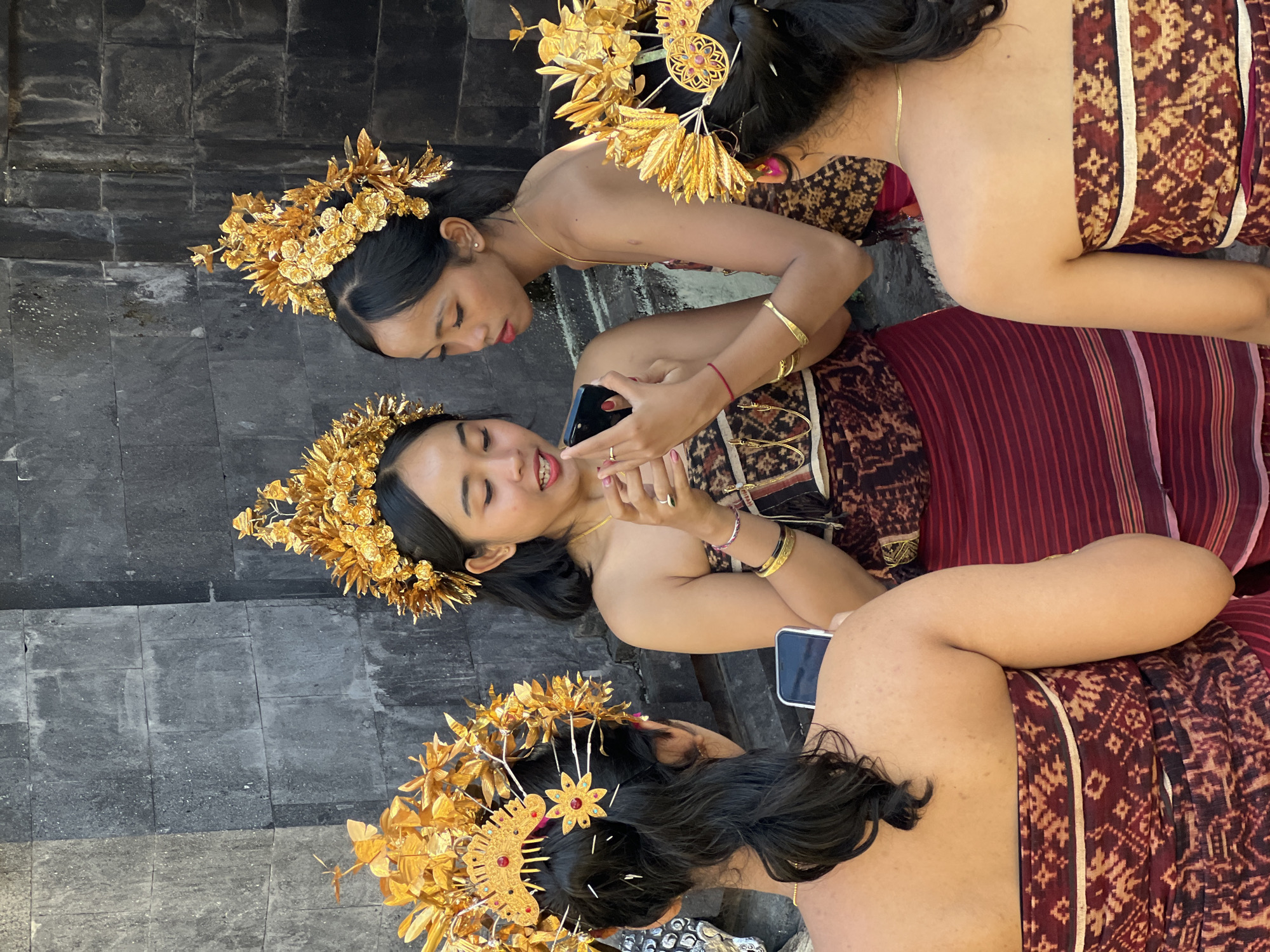 In Manggis, Indonesia, four young women adorned in traditional Balinese attire are gathered outdoors. Their elaborate gold headdresses, resembling intricate crowns with floral motifs and leaf-like accents, are a prominent feature. They are dressed in dark, patterned sarongs and strapless tops, some with metallic gold thread detailing. Two of the women in the center are interacting, one holding up a smartphone and the other looking at it, engaged in conversation. The third woman, facing away, is also holding a smartphone. The fourth woman, also facing away, is partially visible. The backdrop consists of a rough, dark stone wall, suggesting they are in a temple or historical site. The lighting suggests it is daytime, with natural light illuminating the scene.