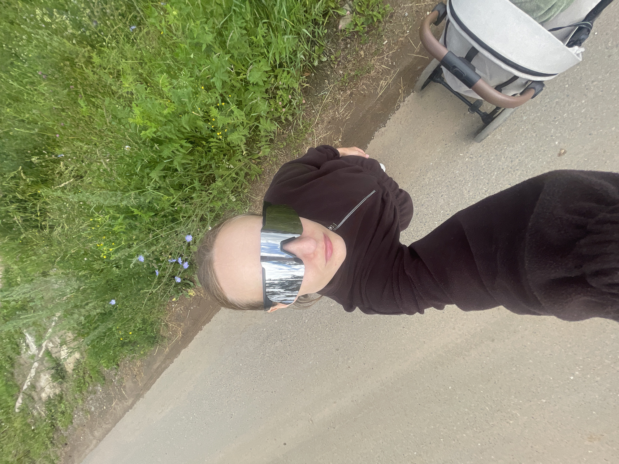 This media file features a person taking a selfie in an outdoor setting in Podshivalovo, Russia. The subject is a young person wearing oversized, mirrored sunglasses and a dark brown hooded jacket with a zipper. The person is outdoors, with a grassy area to their left and a paved path or road to their right. A stroller is partially visible in the background, to the right of the person. The weather appears to be overcast or the lighting is diffused, as the reflections in the sunglasses show a bright, possibly cloudy sky. The overall mood seems casual and relaxed, as if the person is out for a walk or enjoying a leisurely day. The background includes green vegetation with some small flowers. The time of day cannot be precisely determined but appears to be daytime.