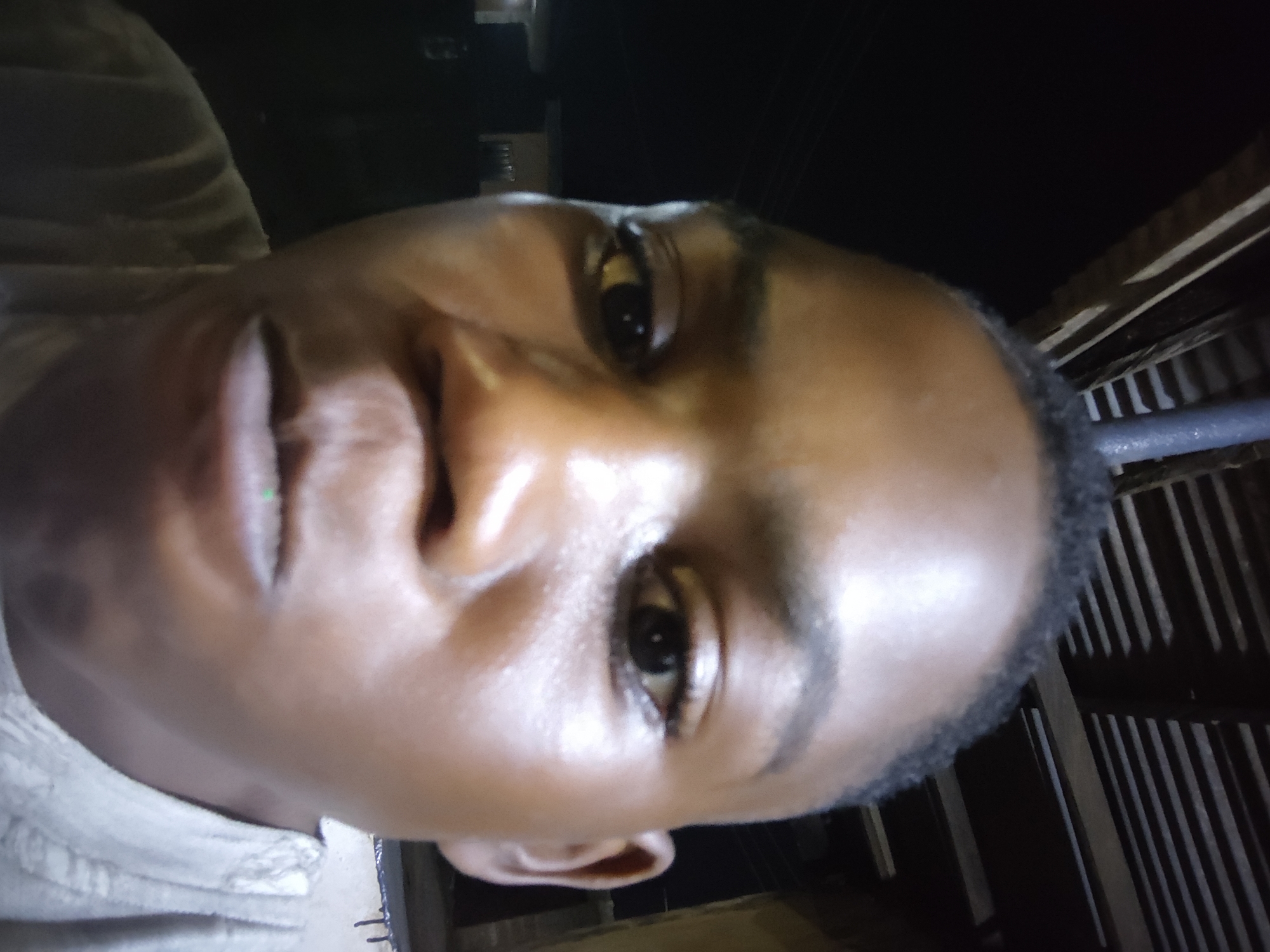 A close-up, low-angle shot of a person's face, presumably male, with brown skin. The person is looking directly at the camera. The background is dark and indistinct, but appears to be a structure with wooden slats. There is no immediate confirmation of the image's location in Ikorodu, Nigeria based solely on the provided picture.