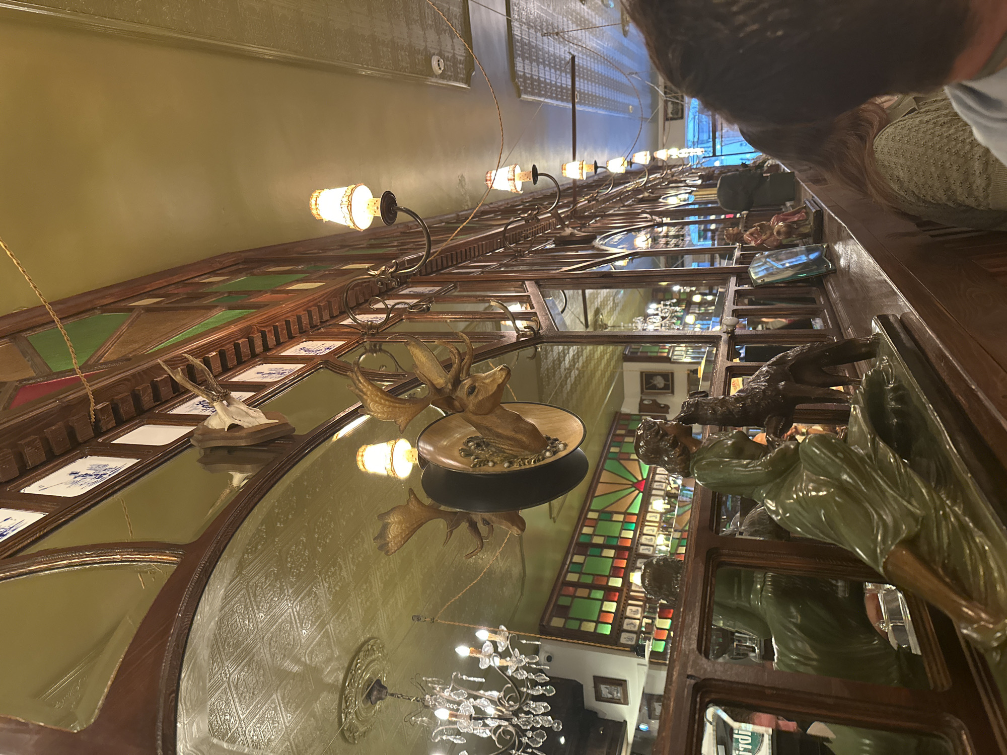 The image depicts the interior of a dimly lit establishment, possibly a bar or restaurant, with a strong emphasis on ornate decor. The scene is dominated by dark wood paneling, mirrored surfaces, and a textured ceiling. A large, mounted deer head with impressive antlers is a focal point, positioned above a stained-glass window featuring geometric patterns in green, red, and yellow. Several wall-mounted lamps with small, fabric-covered shades cast a warm glow. In the foreground, a bronze statue of a reclining woman with a dog is visible, positioned on a wooden ledge with mirrored panels. Reflections in the mirrors show more of the interior, including a chandelier. In the background, people are seated, with one individual's back to the camera, their hair visible. The overall ambiance suggests a vintage or historical setting. While the location is stated to be Bruges, Belgium, there are no specific visual cues within the image to confirm this geographical placement.