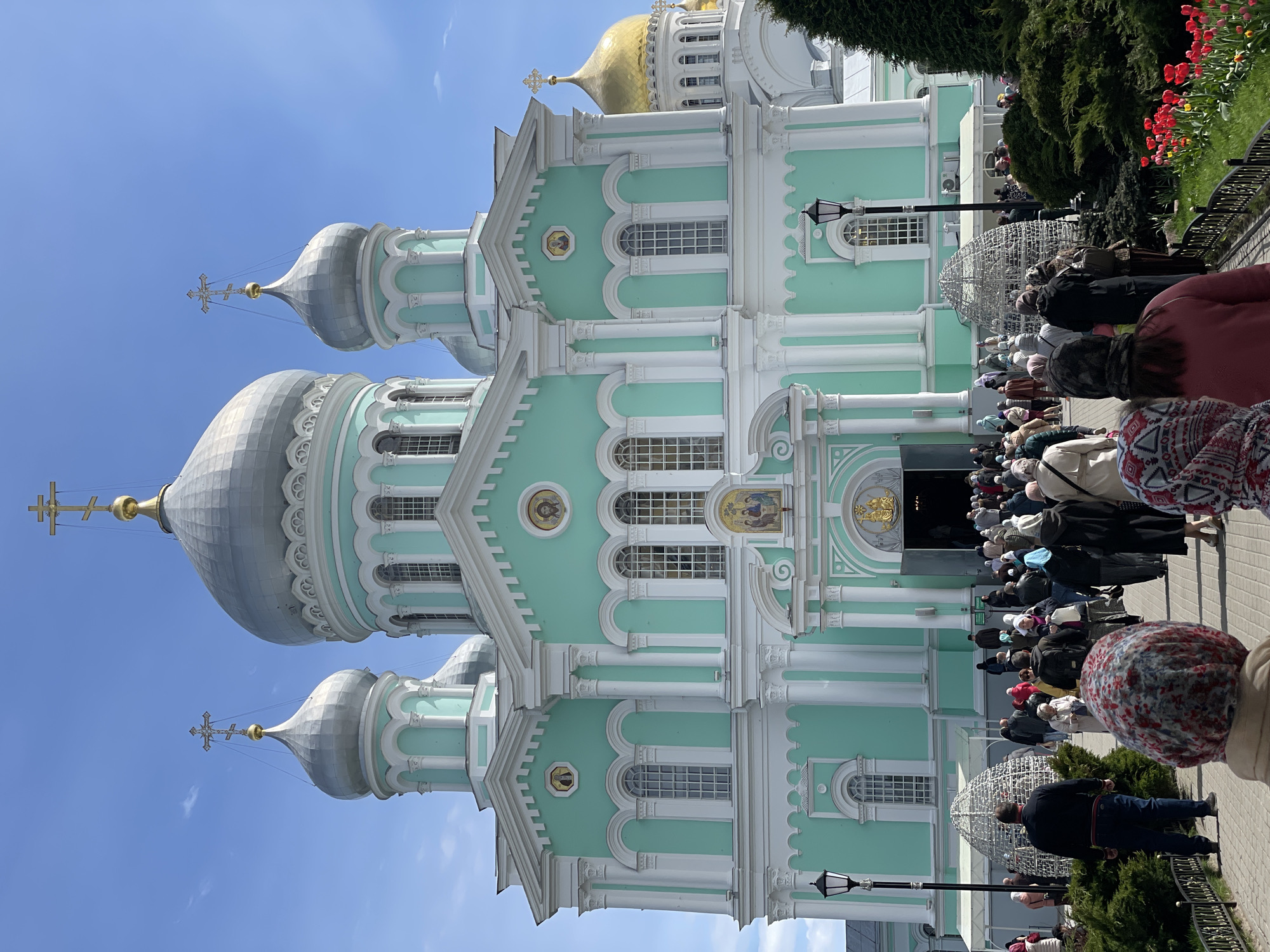 The image shows a tall, light green and white Orthodox church with multiple silver and gold domes, possibly in the city of Unknown, Unknown. A large crowd of people are gathered outside the church entrance. The sky is clear and blue. Flowers are visible on the right side of the frame.