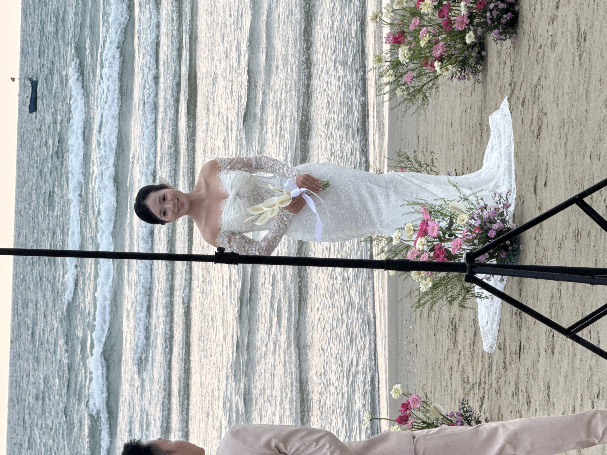 The image shows a bride in a white lace wedding dress holding a bouquet on a beach. Behind her are ocean waves. Floral arrangements frame the scene. A black tripod is visible in the foreground. The setting appears to be a beach wedding, possibly in Đà Nẵng, Vietnam. Another individual wearing a pink suit is partially visible in the bottom of the image.