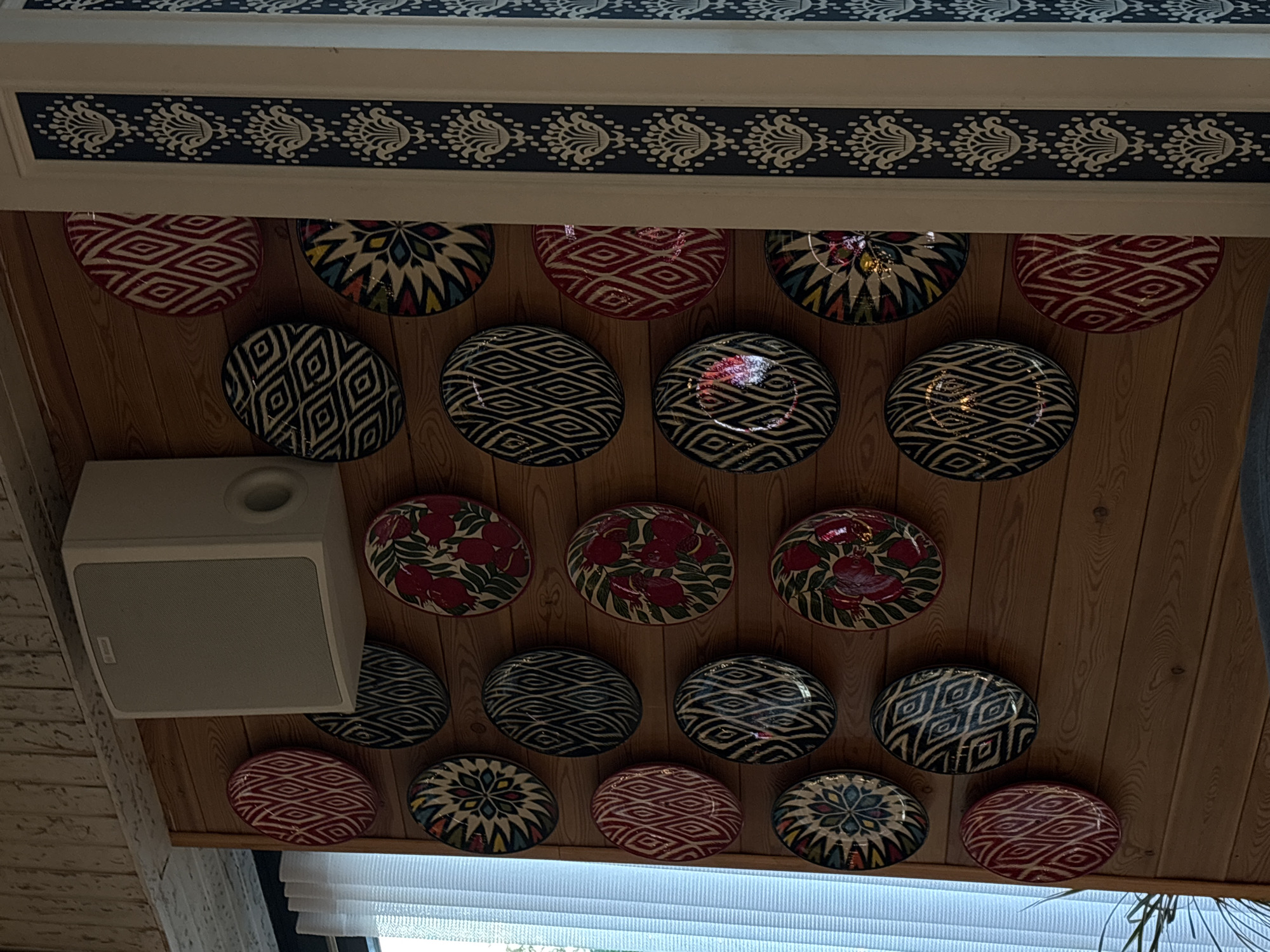 A wall adorned with decorative plates and a speaker is displayed. The wall is made of horizontal wooden planks. On the top of the wall, a white speaker is mounted. Below the speaker, numerous round decorative plates with various patterns are arranged in a grid-like fashion. The plates feature geometric designs in colors like red, black, white, and blue, as well as floral motifs in red and green. To the right of the plates, a vertical decorative trim with a floral pattern in white on a dark blue background is visible. The scene appears to be an interior wall decoration. The location is in the city of Solnechnoye, Russia.