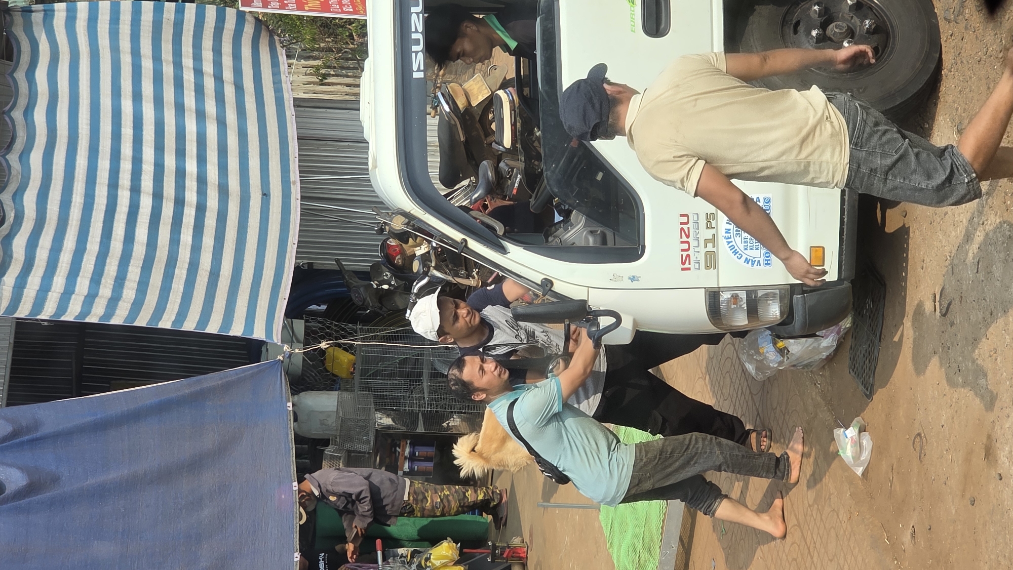 Here is a description of the image:
The image shows a street scene in what appears to be Xuân Long, Vietnam. Several men are working on or around a white Isuzu truck. One man is inside the cabin of the truck, possibly working on the engine. Another man is assisting him from outside, reaching into the engine compartment. A third man is inspecting the rear of the truck, and another stands nearby. There is an assortment of objects nearby, including tools, parts, and a dog. A striped awning provides shade in the background, along with a blue tarp. The ground appears to be paved, and there are some pieces of trash and debris scattered around. The Isuzu truck has an advertisement on the side "ISUZU D-TURA0 91 P5". The location is identified by the OCR on the truck's side "KLBT: H3N VẬN CHUYỂN".