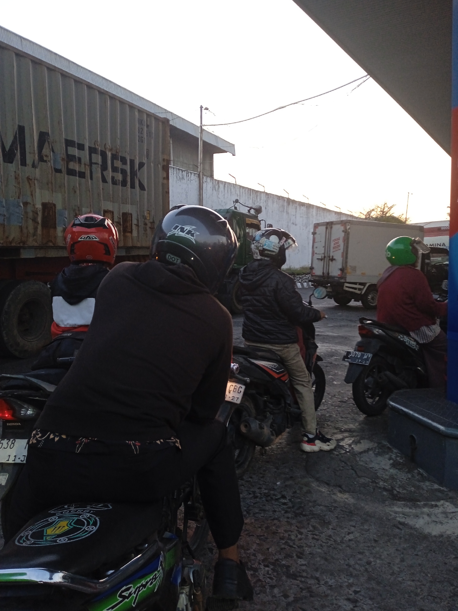 The image shows a group of people on motorcycles at what appears to be a gas station in Semarang, Indonesia. In the foreground, a person wearing a dark brown hoodie and black pants sits on a motorcycle, facing away from the camera. They are wearing a dark helmet with a "INK" logo. To their left, another person on a motorcycle wears a red helmet and a white and black jacket. In the middle ground, two more individuals are on motorcycles. One wears a black jacket and a helmet with various stickers, while the other wears a red jacket and a green helmet. Behind them, a large, weathered shipping container with "MAERSK" written on it is visible. The environment suggests an urban or industrial setting with a paved ground surface and a bright sky.