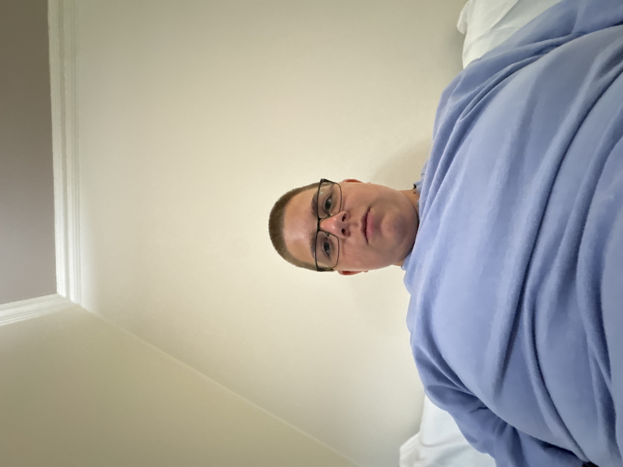 A young man wearing glasses and a light blue sweatshirt is pictured in a close-up, low-angle shot. He is looking directly at the camera with a neutral expression. The background consists of a plain, light-colored wall that meets the ceiling with a white crown molding. There are no discernible landmarks or features that would place this image in Nesebar, Bulgaria. The environment appears to be indoors, possibly a bedroom or living area.
