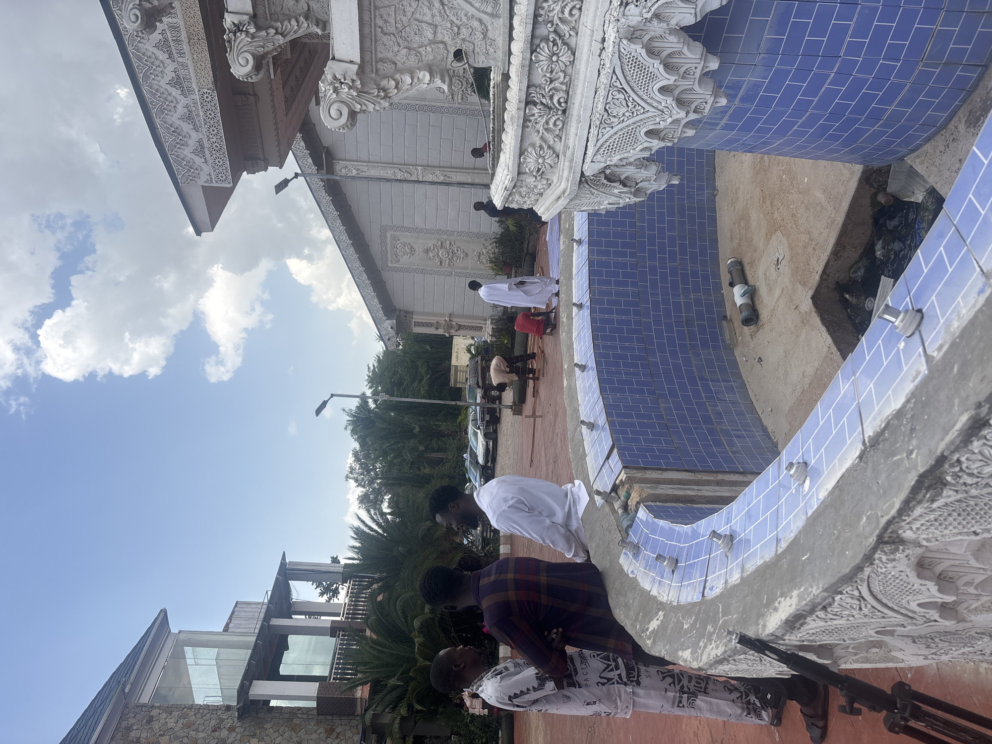 This photograph, taken in Jos, Nigeria, depicts an outdoor scene with several individuals and a partially constructed fountain. The focal point is a circular fountain with blue tiled walls and a concrete rim. Several metal spouts are visible along the rim of the fountain, suggesting it is undergoing installation or renovation. The fountain appears to be empty, with debris visible in a dug-out section within its base.
Several men are present in the scene. Three men are standing in the foreground, engaged in conversation. One man, wearing a white garment with a pattern, is standing to the left. Next to him, a man in a plaid shirt is seated on the fountain's edge. To the right, a man in a white shirt is also standing. Further in the background, other individuals are seen around the fountain and in the surrounding area. Some appear to be working, possibly on the fountain's construction or maintenance, with one individual seen near a rolled-up green mat.
The environment includes elements of a building in the background with distinctive architecture, featuring stone cladding and large glass windows. There are also palm trees and other vegetation, indicating a tropical or subtropical climate. The sky is partly cloudy, with a bright blue expanse. A street light is visible on a pole. In the middle ground, several cars are parked. The overall impression is one of ongoing construction or development in an outdoor setting.