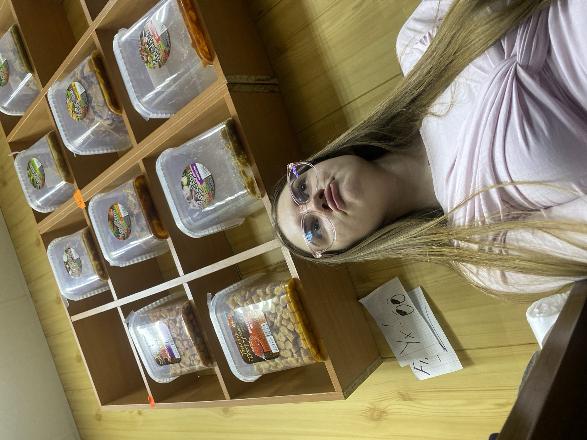 The media file shows a young woman with long blonde hair and glasses lying down in front of a wooden shelf filled with plastic containers. The containers appear to hold food items, with labels visible on some of them. The shelves are organized in a grid pattern, and the overall setting looks like a storage area or a shop. The woman is wearing a light pink, possibly a t-shirt or blouse. Her face is partially visible, and she has a neutral expression. To the right of the woman, there's a piece of paper with a hand-drawn smiley face and some handwritten text in what appears to be Cyrillic script. The lighting suggests it might be daytime. The location is identified as Дьяково, Russia.