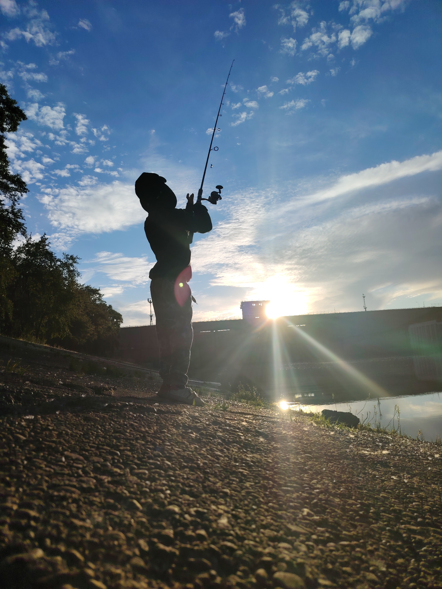 A silhouetted child stands on a gravelly bank, holding a fishing rod and looking up at the sky. The sun is setting, casting a warm glow and lens flares across the scene. In the background, a dam structure is visible, with water reflecting the sky and the sun. Trees line the left side of the image, and the sky is filled with scattered clouds. The scene appears to be taking place near a body of water, possibly a reservoir or river. The location is identified as Chaikovsky, Russia.