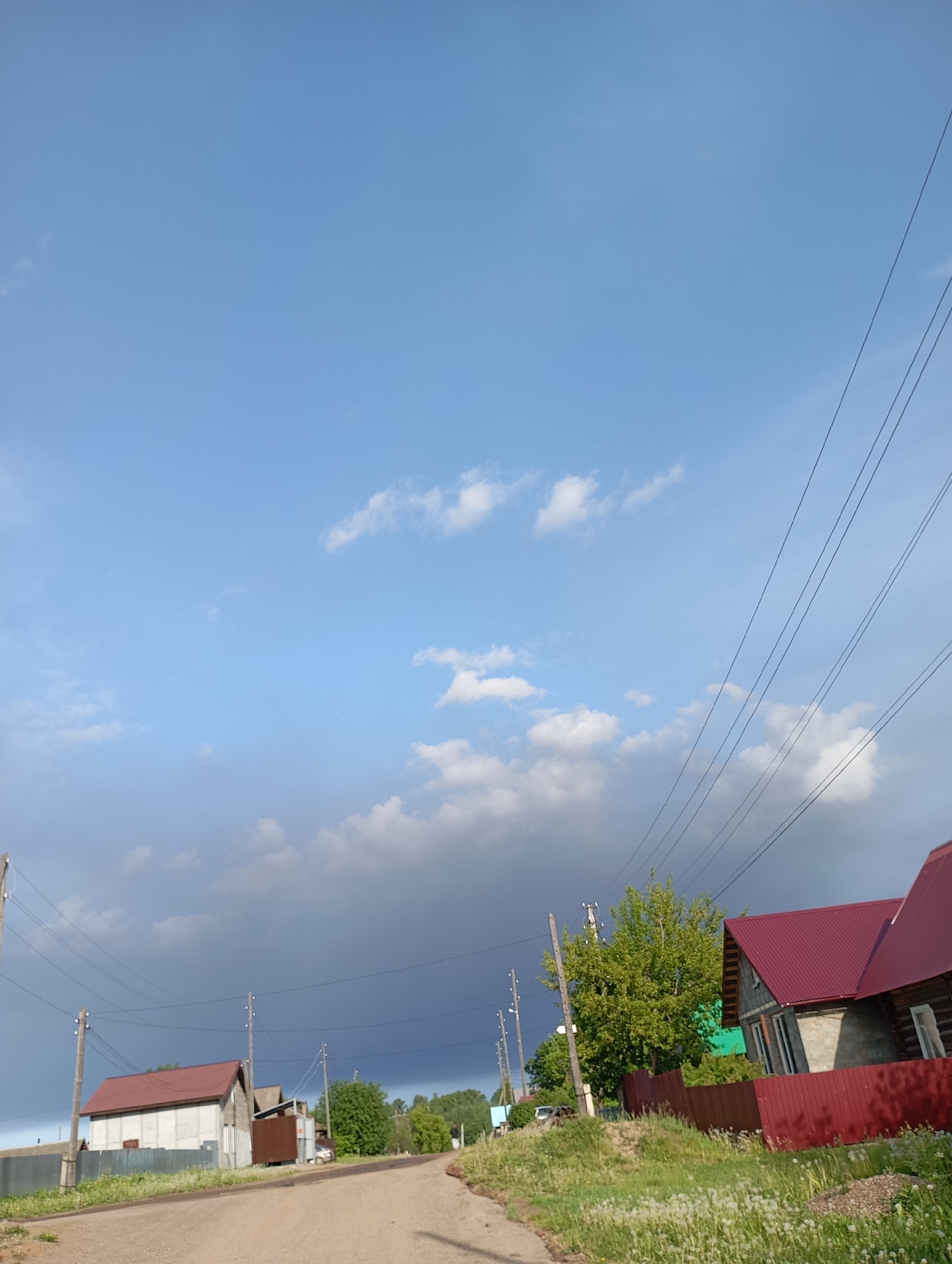 The image depicts a rural street in Фоки, Russia. A dirt road stretches into the distance, lined with houses. The houses are primarily made of wood or concrete and have red metal roofs. Power lines run along the side of the road. Vegetation is present on the sides of the road. A dark storm cloud hangs in the sky above.
