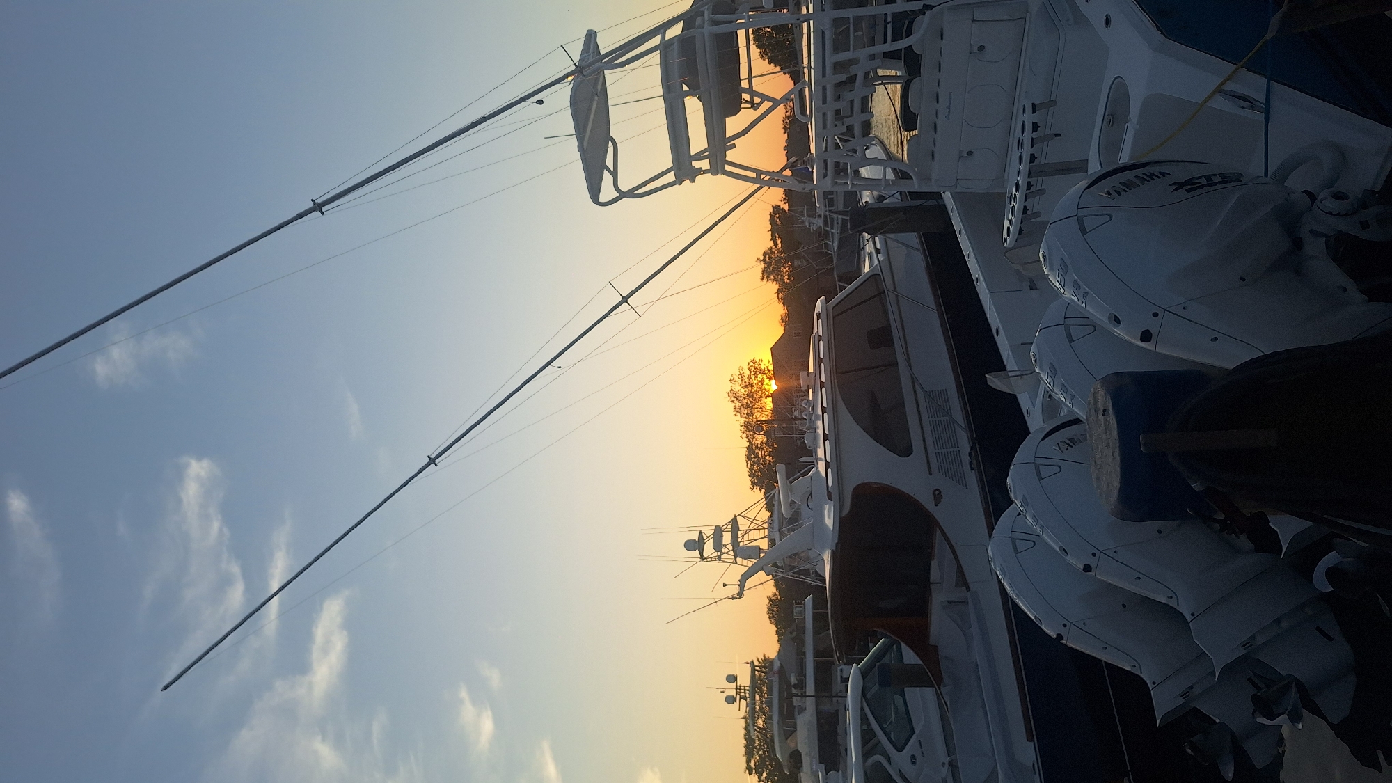 The image depicts a serene sunset scene at a harbor in Nantucket, Massachusetts, USA. Several large yachts and boats are docked, with their masts and rigging silhouetted against the warm hues of the setting sun. The sky is a gradient of blue and orange, with wispy clouds adding texture. In the foreground, the powerful outboard engines of a boat are visible, hinting at the recreational or fishing purpose of the vessels. The overall atmosphere is peaceful and picturesque, capturing the beauty of a coastal sunset.