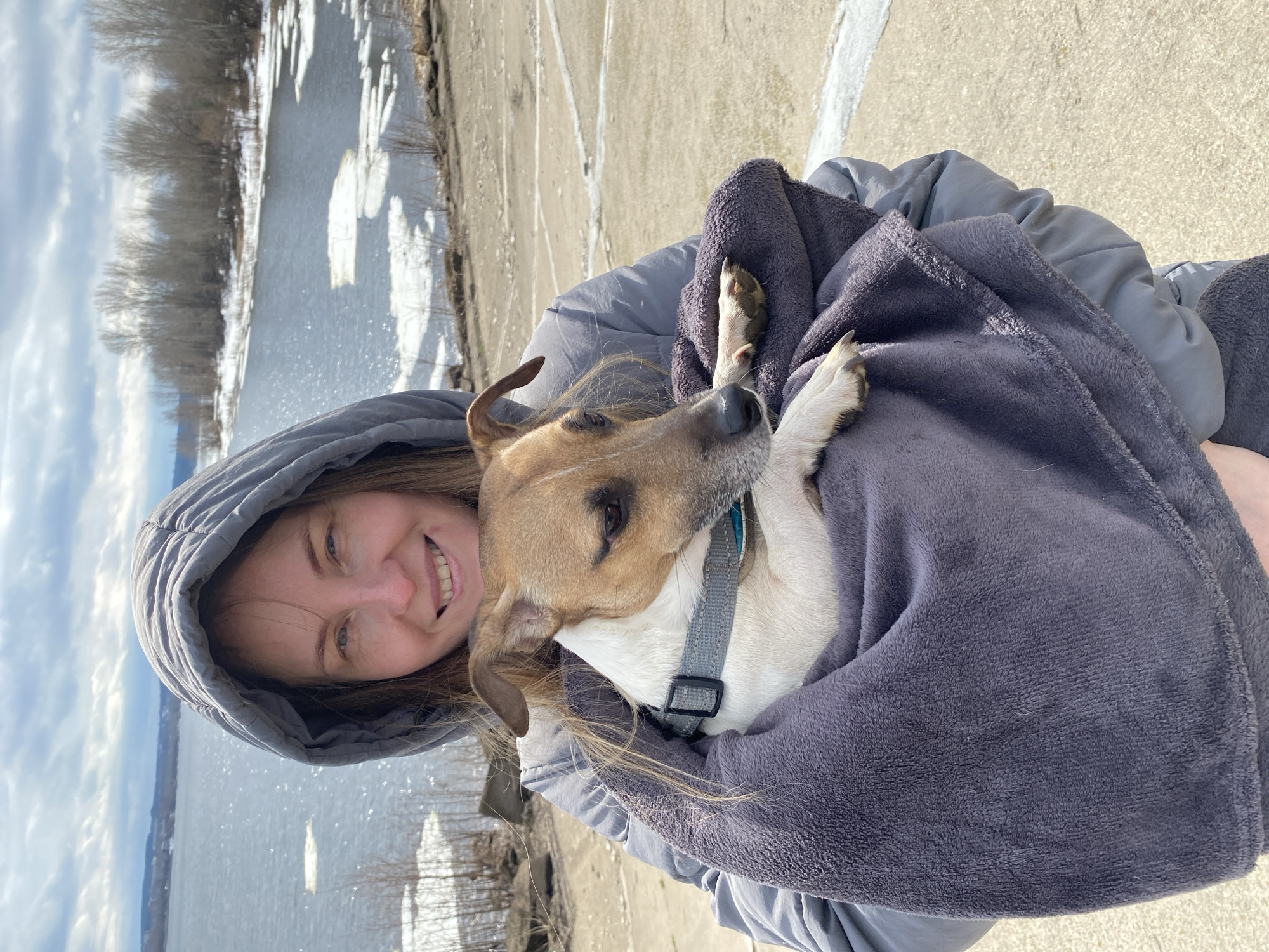 A woman in a hooded gray jacket is holding a dog wrapped in a gray blanket. The dog is light brown and white, and wears a gray collar. The woman is smiling. Behind them is a river partially covered with ice, with trees lining the opposite bank. The location is likely in the city of Chaikovsky, Russia. A concrete surface is visible near the woman and dog.