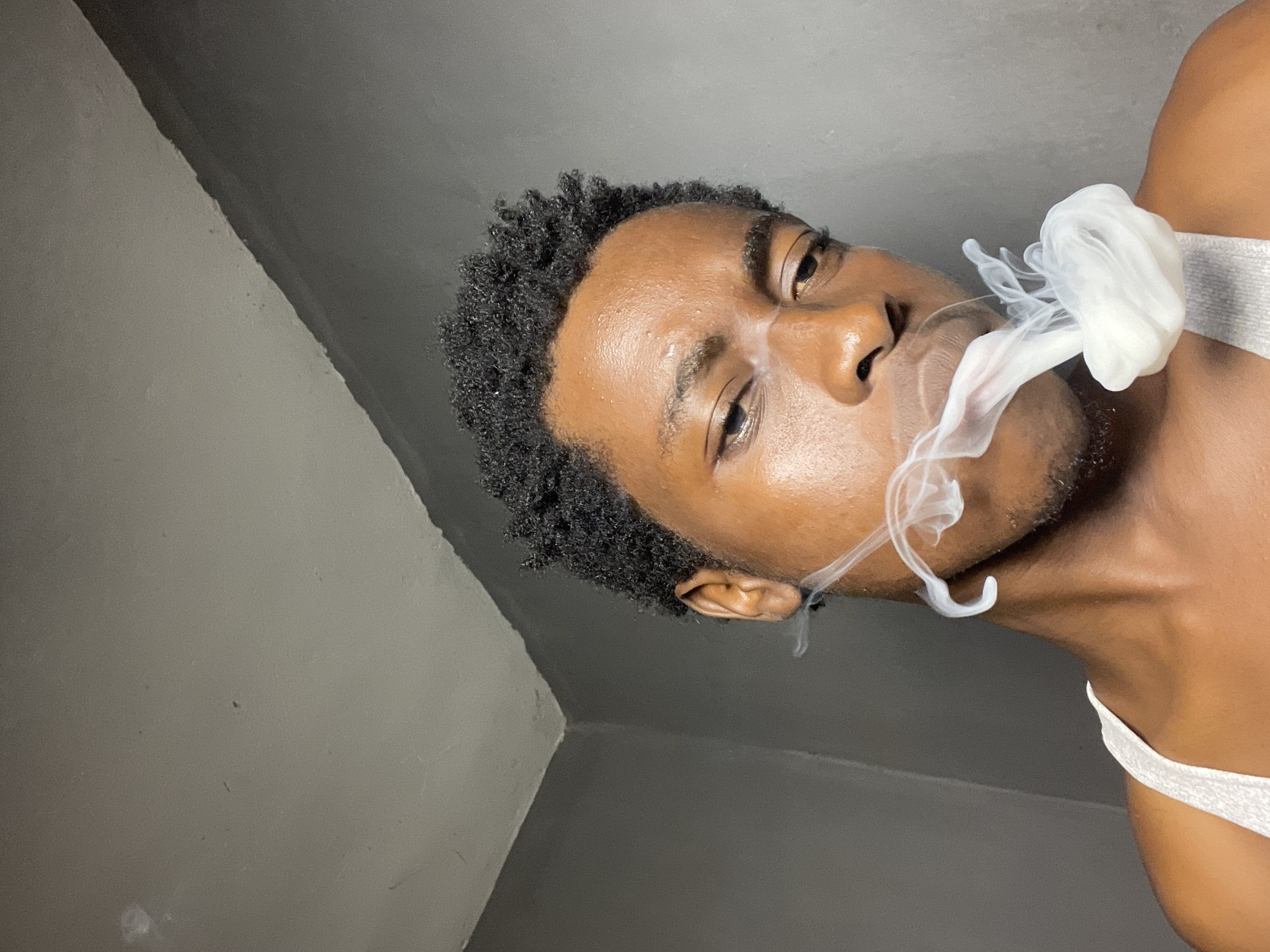 A close-up, head-and-shoulders shot captures a young Black man exhaling a plume of white smoke. He is wearing a white tank top and has short, dark, curly hair. The background consists of a grey, textured wall with a visible seam where the wall meets the ceiling, indicating an indoor setting. The lighting is dim, with a spotlight seemingly coming from the left, casting shadows on the man's face and accentuating the texture of his skin. The composition is tight, focusing on the man's face and the smoke. The image does not provide any specific visual cues that can definitively place the location within Nkpor, Nigeria.
