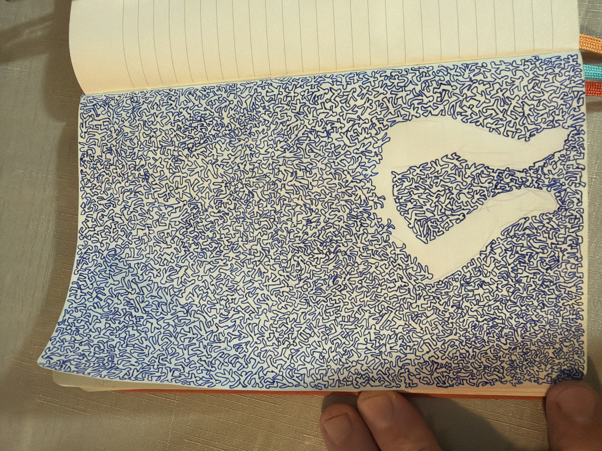 The image shows an open notebook with a drawing on the left page. The drawing is made with blue ink and consists of a dense pattern of small, abstract figures that resemble stylized human shapes. These figures are interconnected and fill the majority of the page. In the center of the drawing, there is a large negative space in the shape of two figures embracing. The right page of the notebook is blank and lined. The notebook is placed on a textured surface, possibly a table. A person's fingers are visible on the left edge of the image, holding the notebook. The image does not contain any information or visual cues that would allow for the identification of a location in Even Yehuda, Israel.