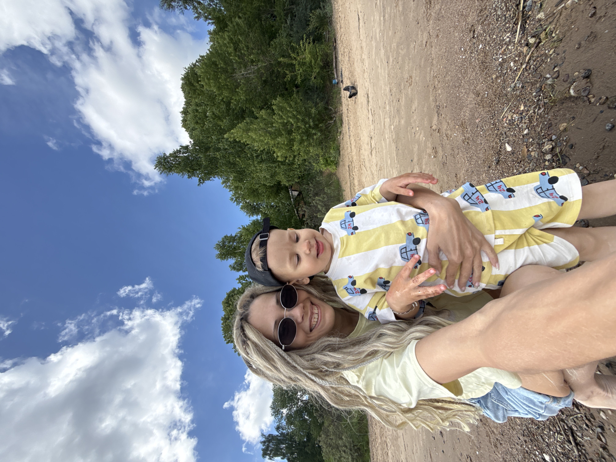 A selfie-style portrait features a smiling woman holding a young child outdoors on a sunny day. The woman, positioned closer to the camera, has long, wavy blonde dreadlocks or braids, round dark sunglasses, a light yellow t-shirt, and blue denim shorts. She smiles broadly. The child, held in her arms, has light blonde hair, wears a black baseball cap backwards, and a yellow and white striped shirt with blue truck illustrations. The child's eyes are closed in a joyful expression, and he appears to be smiling or laughing.
The background shows a vibrant blue sky with several large, fluffy white clouds. Behind the subjects and stretching across the midground is a dense area of lush green trees or bushes. The ground beneath them is a sandy or light brown dirt, with patches of darker soil and scattered small rocks and pebbles, suggesting a natural outdoor environment like a beach or riverbank. The bright lighting indicates it is daytime. Both individuals appear happy and content. No prominent text is visible, although small details are part of the truck patterns on the child's shirt.