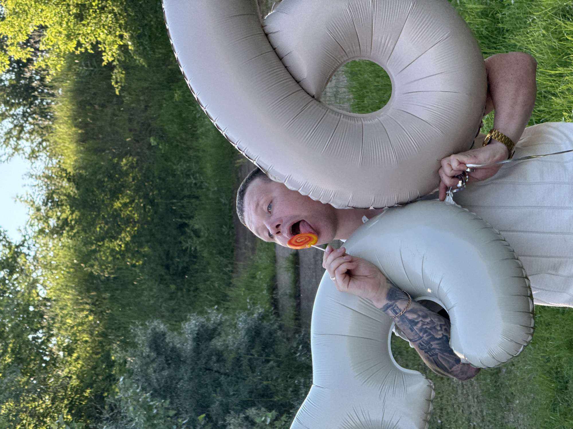 The image shows a man with a lollipop in his mouth, holding two large, light-colored balloons that form the number "16". He is wearing a light-colored shirt and a gold watch. He has tattoos on his left arm. The background consists of lush green trees and grass, suggesting an outdoor setting. The lighting indicates it is daytime. The image does not contain any specific landmarks or indicators to pinpoint a location within Chaikovsky, Russia.