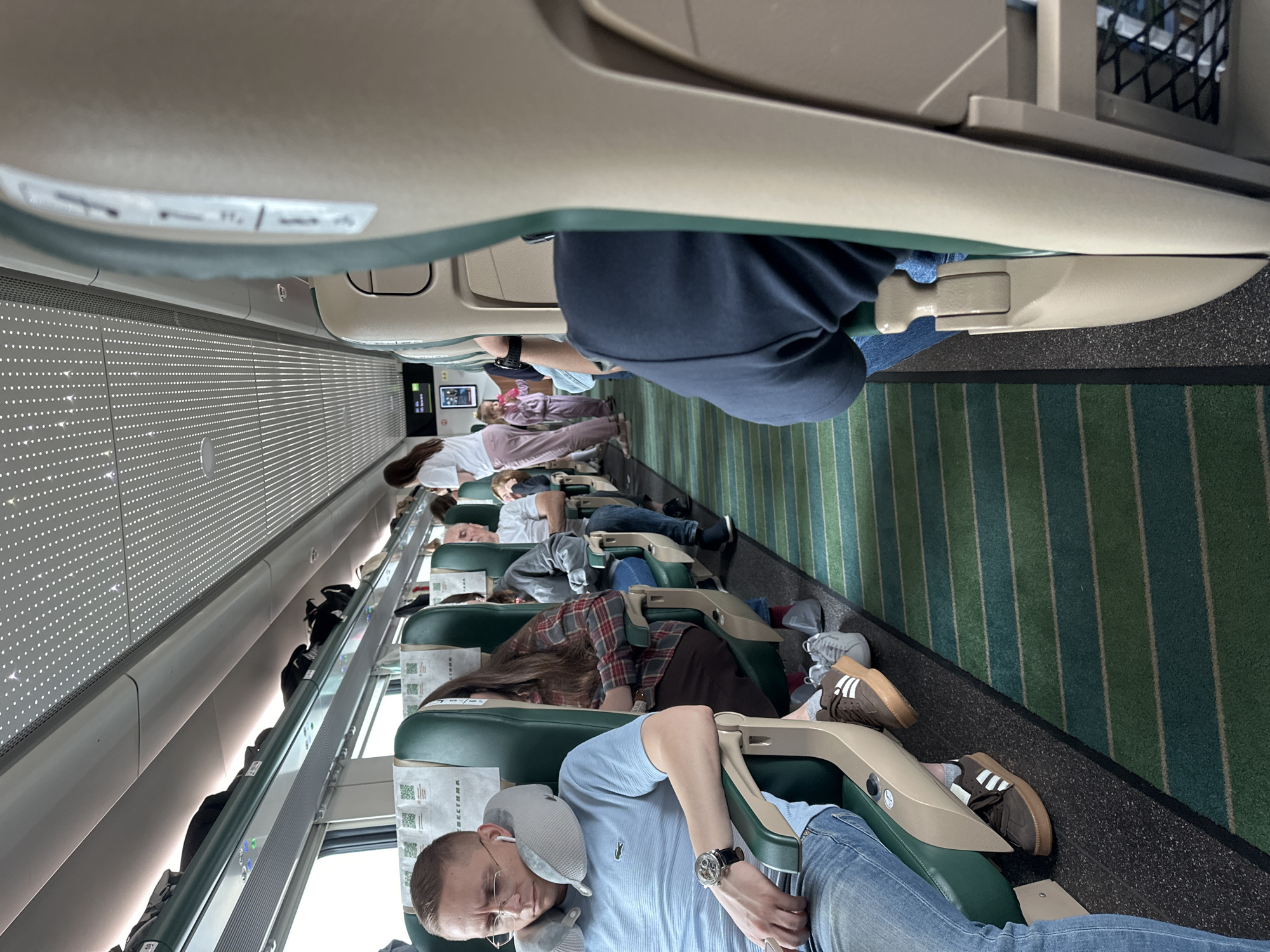 The image depicts the interior of a train carriage. In the foreground, a man wearing a light blue t-shirt and jeans is seated, leaning his head on a gray neck pillow. He has earbuds in and is wearing glasses and a wristwatch. To his right, a woman in a plaid shirt is seated, and further down the aisle, a man in a gray hoodie and jeans is visible. At the end of the carriage, a woman in a white shirt and pink pants and a young girl in a pink tracksuit are standing. Luggage is stored in the overhead compartments. The train has green and blue striped carpeting on the floor and green seats with light beige headrests and armrests. The ceiling features a patterned design with lights. The overall scene suggests a passenger train journey. The image was taken inside the Sobinka train station, located in Sobinka, Russia.