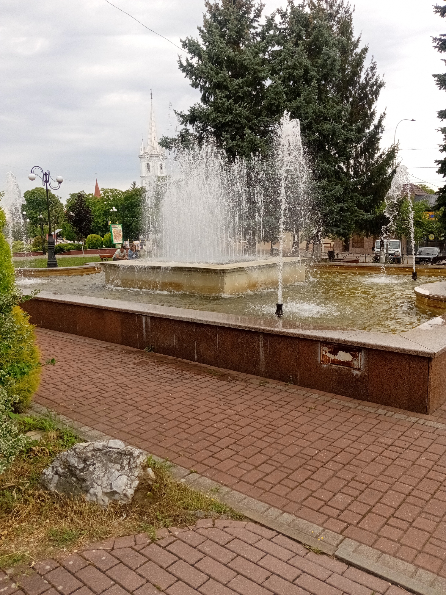 The image depicts a fountain in a park area in Tiachiv, Ukraine. The fountain is the central element, with water jets spraying upwards from a square basin. The basin is made of brown granite-like material. To the left of the fountain, a street lamp with two globes is visible. In the background, there are trees, including large coniferous trees, and a white church spire with intricate architectural details. A few people are seated near the fountain, on a bench. The foreground is paved with reddish-brown brick pavers, and there is some grass and a large grey rock to the left of the path. The sky is overcast. The overall scene suggests a public recreational space in the city.