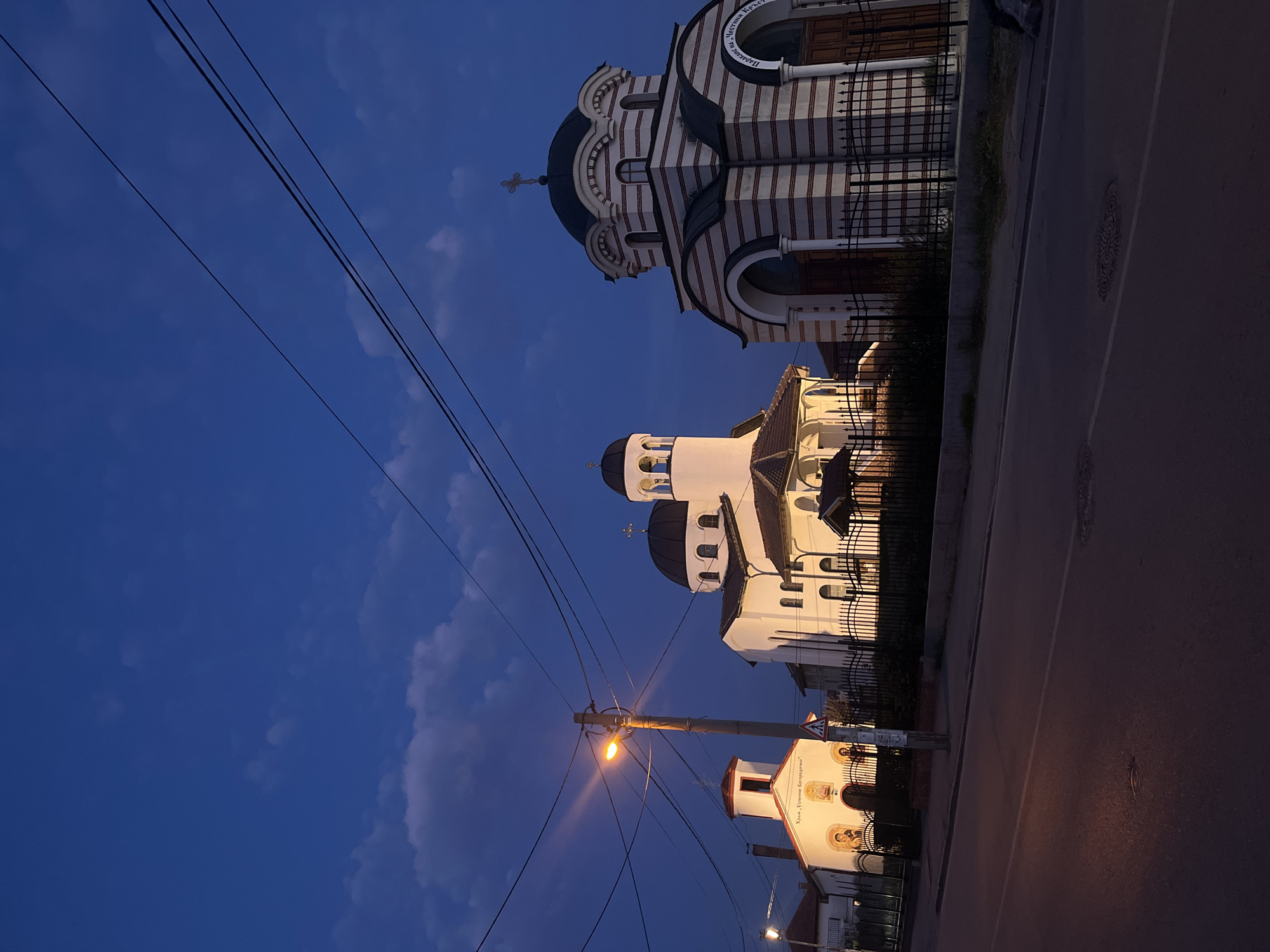 The image depicts a street scene in Sofia, Bulgaria, at twilight. Three church buildings are visible: one striped brown and white, one white with black domes, and a smaller white building with religious paintings on its facade. A street runs along the right side of the frame, and overhead power lines crisscross the sky. A streetlight illuminates the scene. The sky is a deep blue with some clouds.