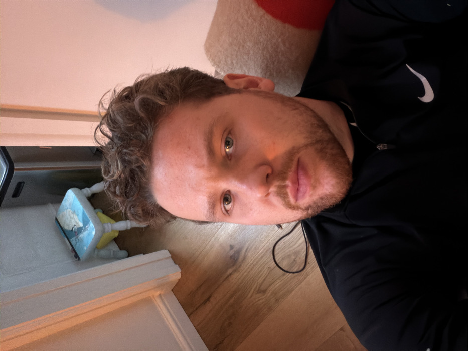A male individual, estimated to be in his mid-20s to early 30s, with fair skin, light-colored eyes, light brown curly hair, and a short brown beard/stubble, is captured in a close-up, self-portrait-style photograph taken within a domestic interior in London, United Kingdom. He is wearing a black long-sleeved quarter-zip top featuring a white Nike swoosh logo on the right chest. He directly faces the camera, positioned slightly below eye level.
The background reveals elements of a residential space. To the left of the subject, a black cable is coiled on a light brown wooden floor. Further into the background, a white baseboard runs along a light-colored wall, leading to a partial view of a white doorframe. A small, light blue wooden step stool with white legs and a graphic design on its surface is positioned near the wall. Behind the stool, a stainless steel trash can with a black lid is visible. To the right of the subject, a portion of a beige textured cushion or blanket with a red section is present. The scene is illuminated by a warm, reddish-orange light. There are no other individuals or active interactions present.