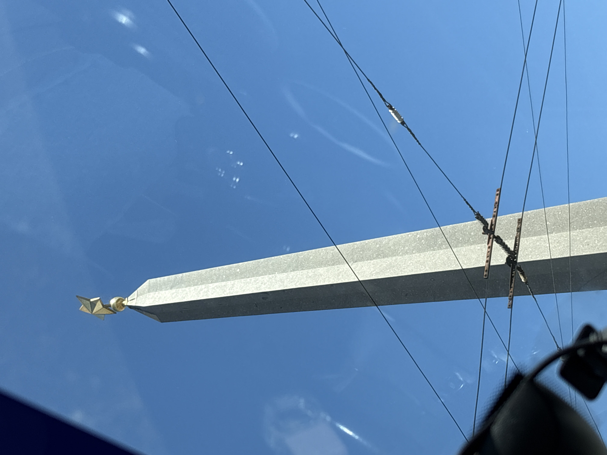This image shows a tall, grey, four-sided obelisk topped with a gold star, set against a bright blue sky. Several dark wires crisscross in front of the obelisk, some of them supported by rusty metal fixtures. The perspective suggests the photo was taken from a low angle, possibly from a vehicle, as indicated by a dark, blurry object in the lower left corner, which could be part of a car's interior. The lighting suggests it is daytime, with some reflections visible on what appears to be a window. The obelisk is likely the Narva Triumphal Arch, a monument located in St. Petersburg, Russia.