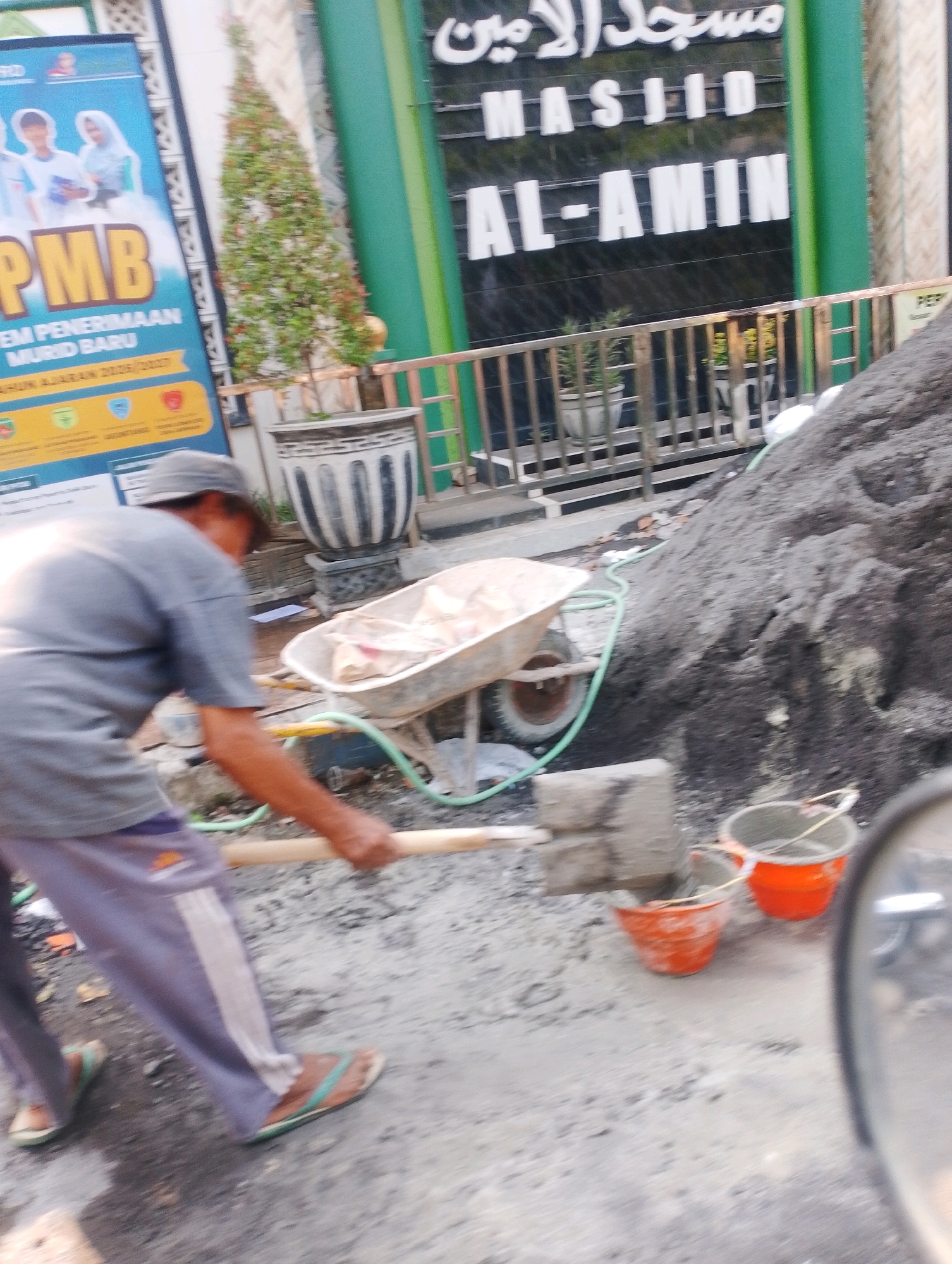 A person is shoveling sand into a wheelbarrow at a construction site. The sand pile is large and dark gray. The wheelbarrow is metallic gray and has a light-colored bag inside. There are two orange buckets and one gray bucket nearby. The person is wearing a gray shirt, gray hat, and light purple pants with a white stripe. They are also wearing brown flip-flops. In the background, there is a building with Arabic writing on it that says "Masjid Al-Amin". There is a green fence and some potted plants. There is also a blue banner with images of people and text about school enrollment. The scene is outdoors and appears to be in a city. The Arabic writing on the building and the style of the banner suggest the location is in Indonesia, likely Probolinggo, Indonesia, given the context of the prompt.