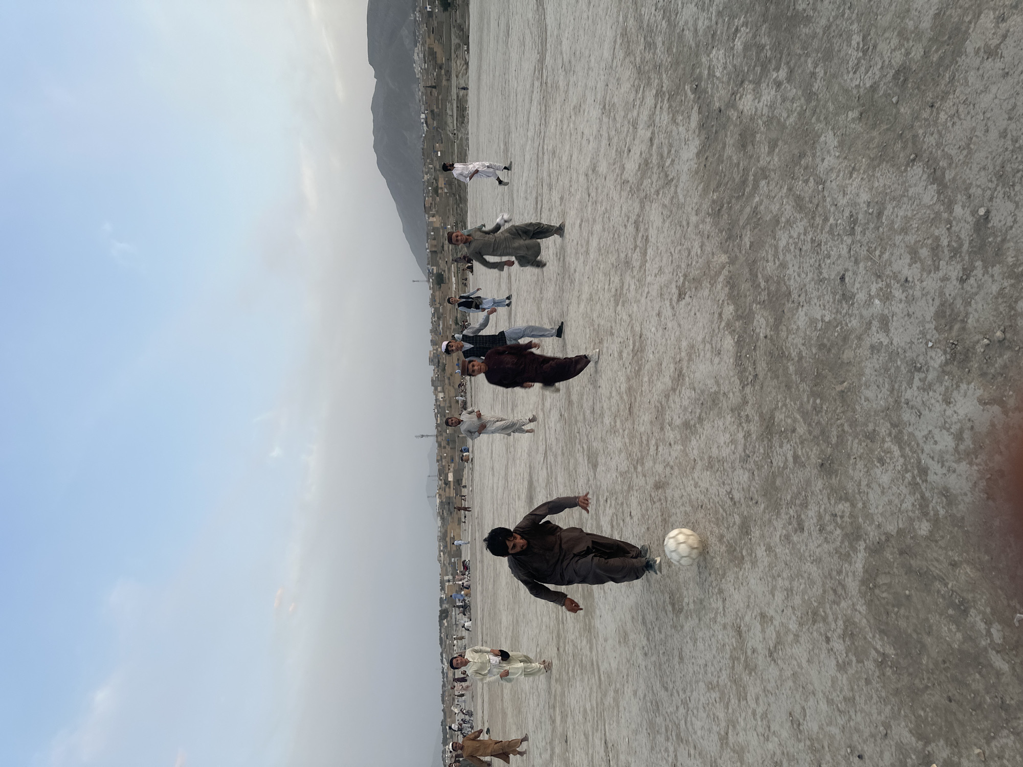 People play soccer on a dirt field outside of Quetta City Tehsil, Pakistan. A row of buildings is on the edge of the field, with mountains in the background. A soccer ball is visible on the field, with one person kicking the ball.