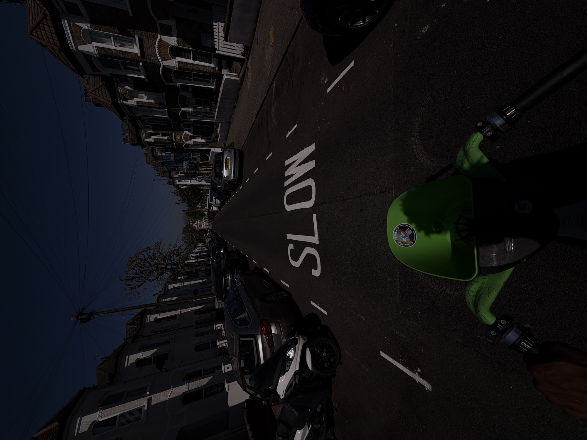 Captured from a tilted perspective, the image showcases an urban street scene in what appears to be London, United Kingdom. The street is lined with rows of townhouses and various parked vehicles. The word "SLOW" is painted prominently on the asphalt roadway. In the foreground, there is a visible segment of a green object, presumably a scooter or similar vehicle. Power lines intersect the sky in the upper left. The scene is bathed in sunlight, creating harsh shadows.