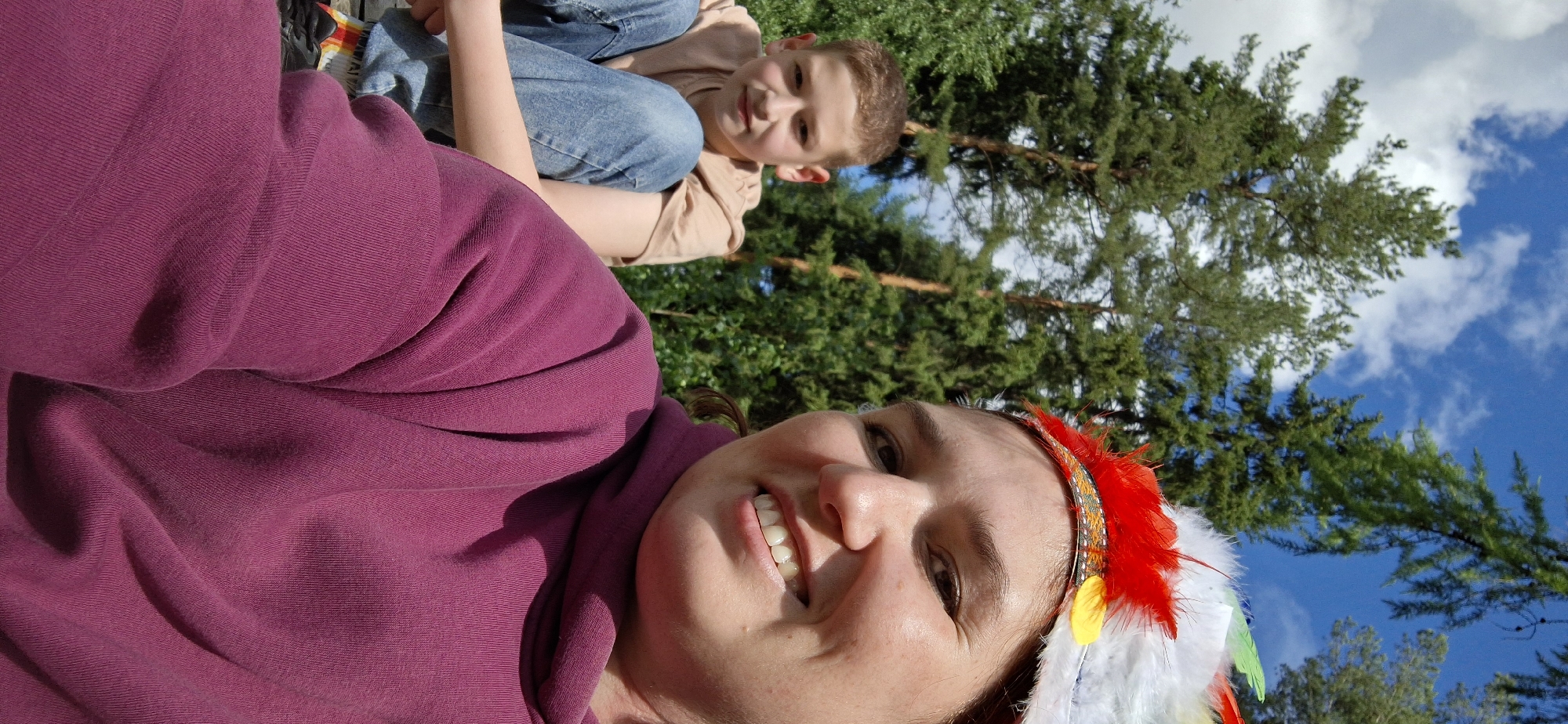 The image shows a woman and a child outdoors, possibly in a park or natural area in or near Прикамский, Russia. The woman is in the foreground, wearing a maroon-colored sweatshirt or hoodie, smiling. She is also wearing a feathered headdress with red, yellow, and white feathers. Behind her, to the right, is a young child. The child is seated, wearing a tan shirt and denim jeans. They are looking toward the camera. The background features lush green trees, a blue sky, and white clouds. The image is taken at an unusual angle, slightly tilted.