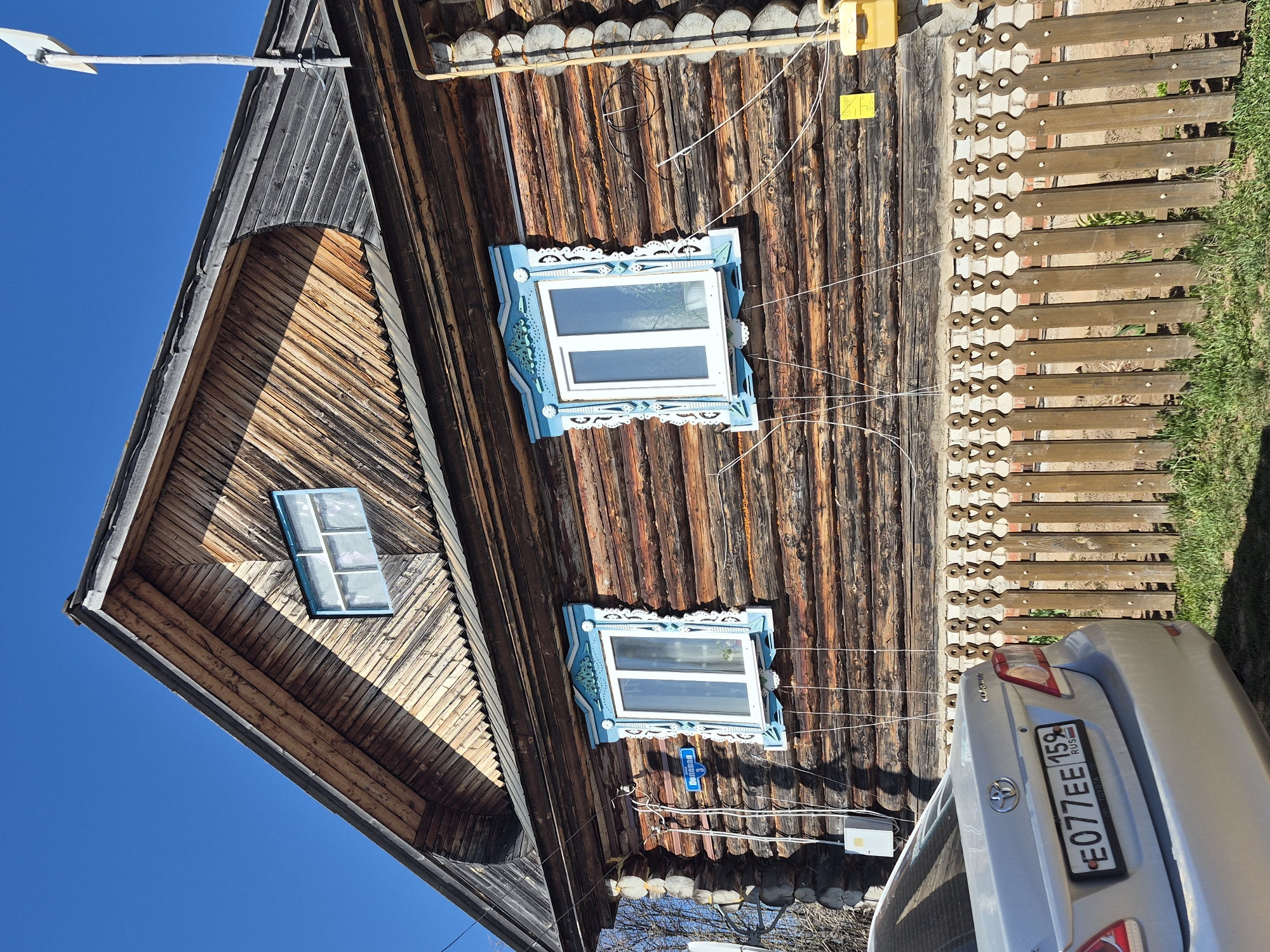 The image shows a wooden house with blue window frames in Дойная, Russia. A silver Toyota car with the license plate E077EE 159 RUS is parked in front of a wooden fence. The sky is blue.